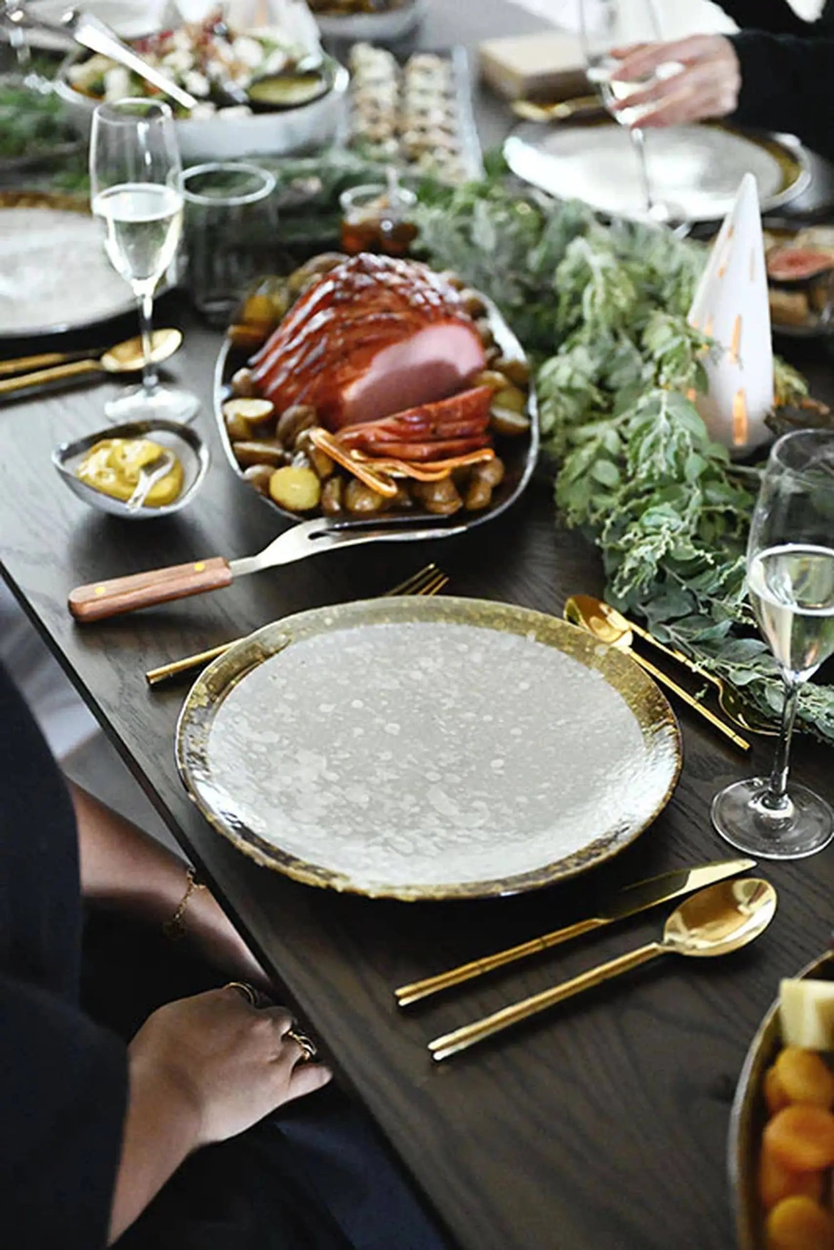 A festive table set with gold cutlery, Kra Sanctuary Satay Free-Form Dinner Plates, wine glasses, greenery decor, and a platter of roasted meat with potatoes in the background.