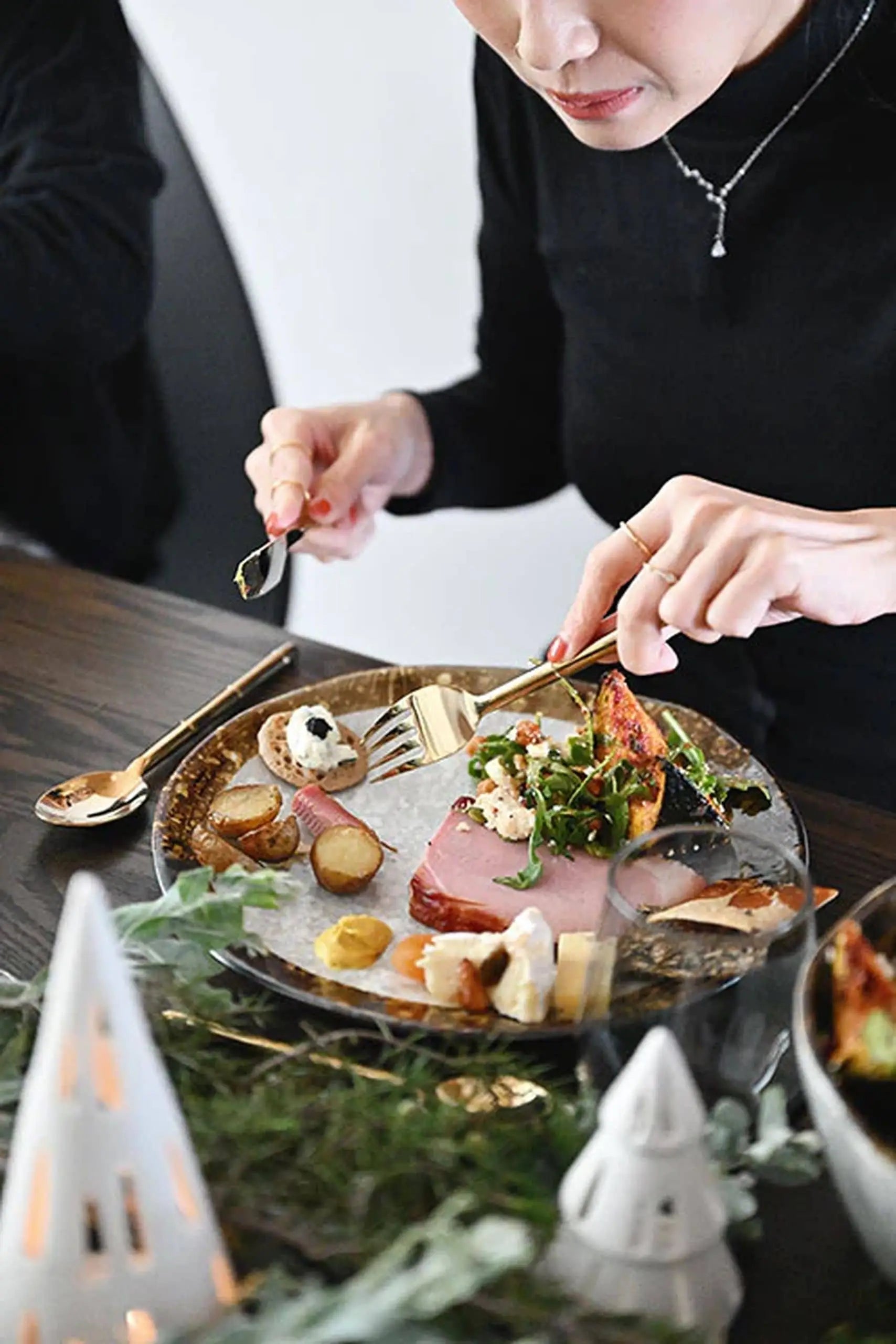 A person in a black top slices into a gourmet meal on the Kra Sanctuary Satay Free-Form Dinner Plate by Kra Sanctuary, with holiday ornaments and earthy plates featuring an iridescent border set atop a dark wooden table.