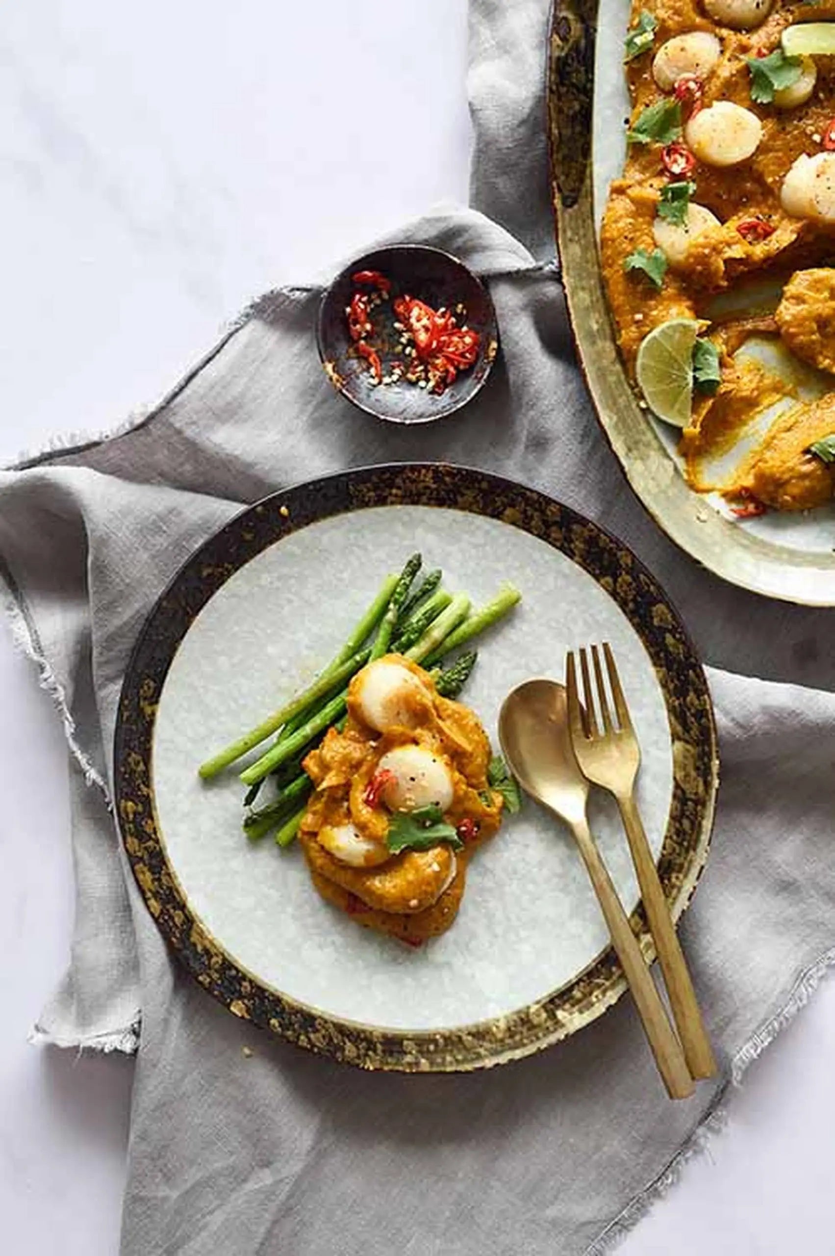 Scallops on golden curry sauce with asparagus are served on the Kra Sanctuary Satay Free-Form Dinner Plate by Kra Sanctuary, accompanied by a gold fork and spoon. A side dish with sauce and sliced chili sits nearby on a gray napkin.