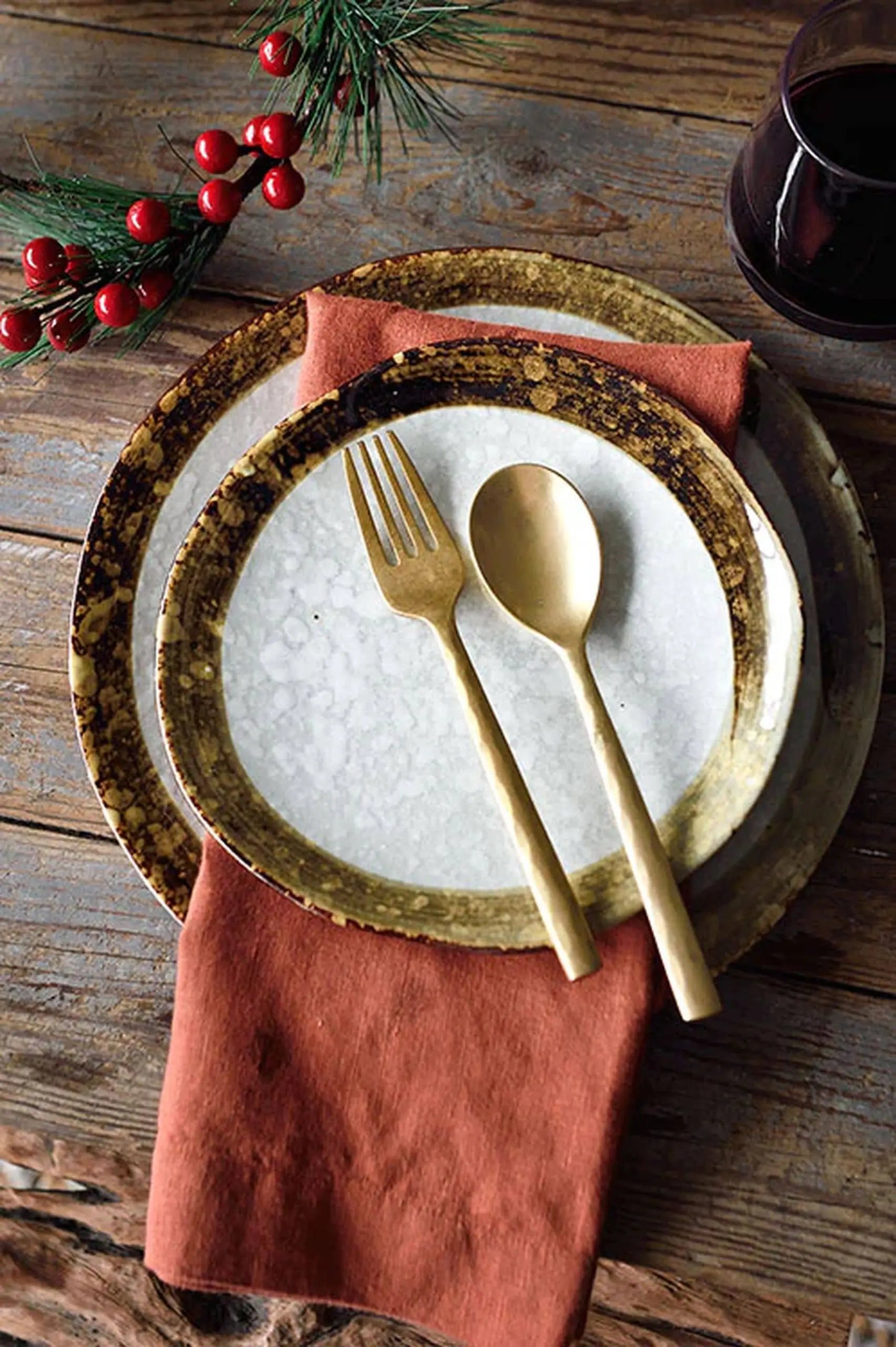 A rustic table setting features Kra Sanctuary Satay Free-Form Side Plates, gold utensils on a burnt orange napkin, and a sprig of greenery with red berries atop a wooden table. A glass of dark liquid sits in the upper right corner.