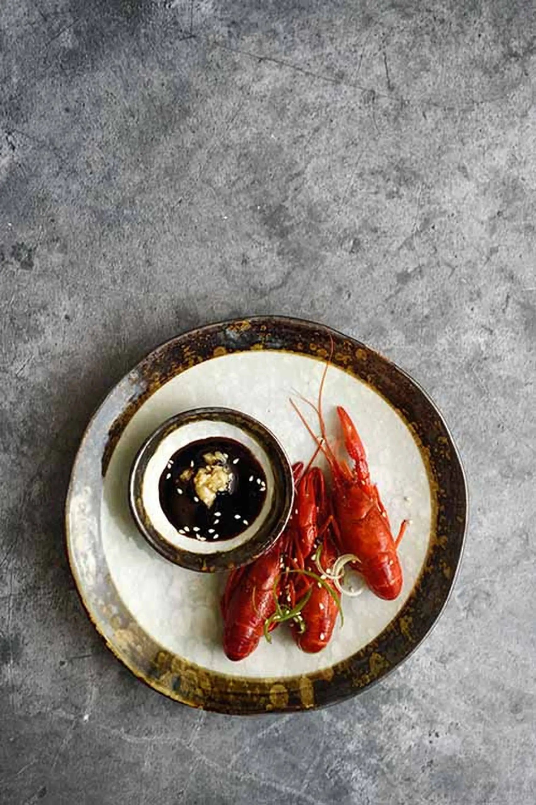 Two cooked crayfish are served on the Kra Sanctuary Satay Free-Form Side Plate, accompanied by a bowl of dark dipping sauce with sesame seeds and garlic, all set on a textured gray surface for an elevated dining experience.