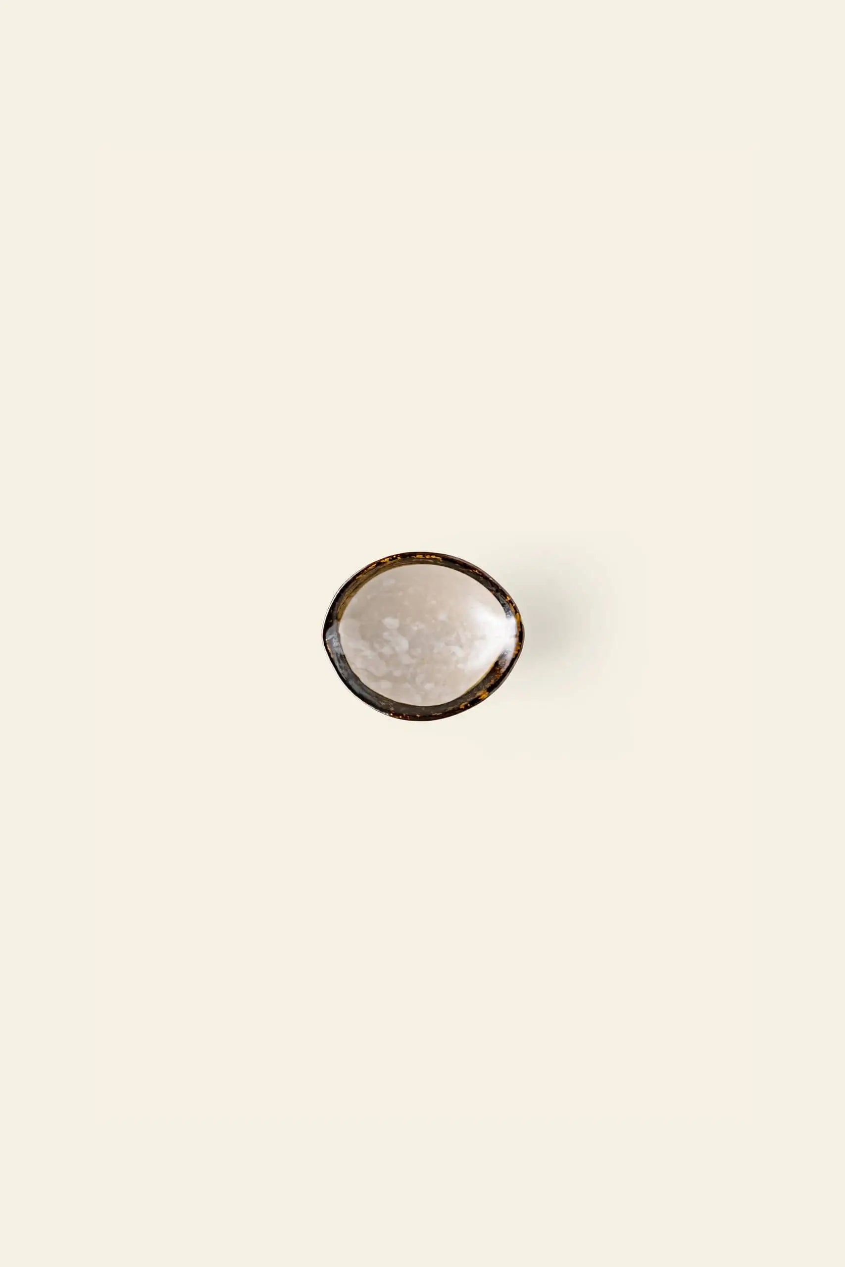 The Kra Sanctuary Satay Shell Dipping Dish by Kra Sanctuary, featuring a gently textured surface and an iridescent edge, is centered on a plain, off-white background.