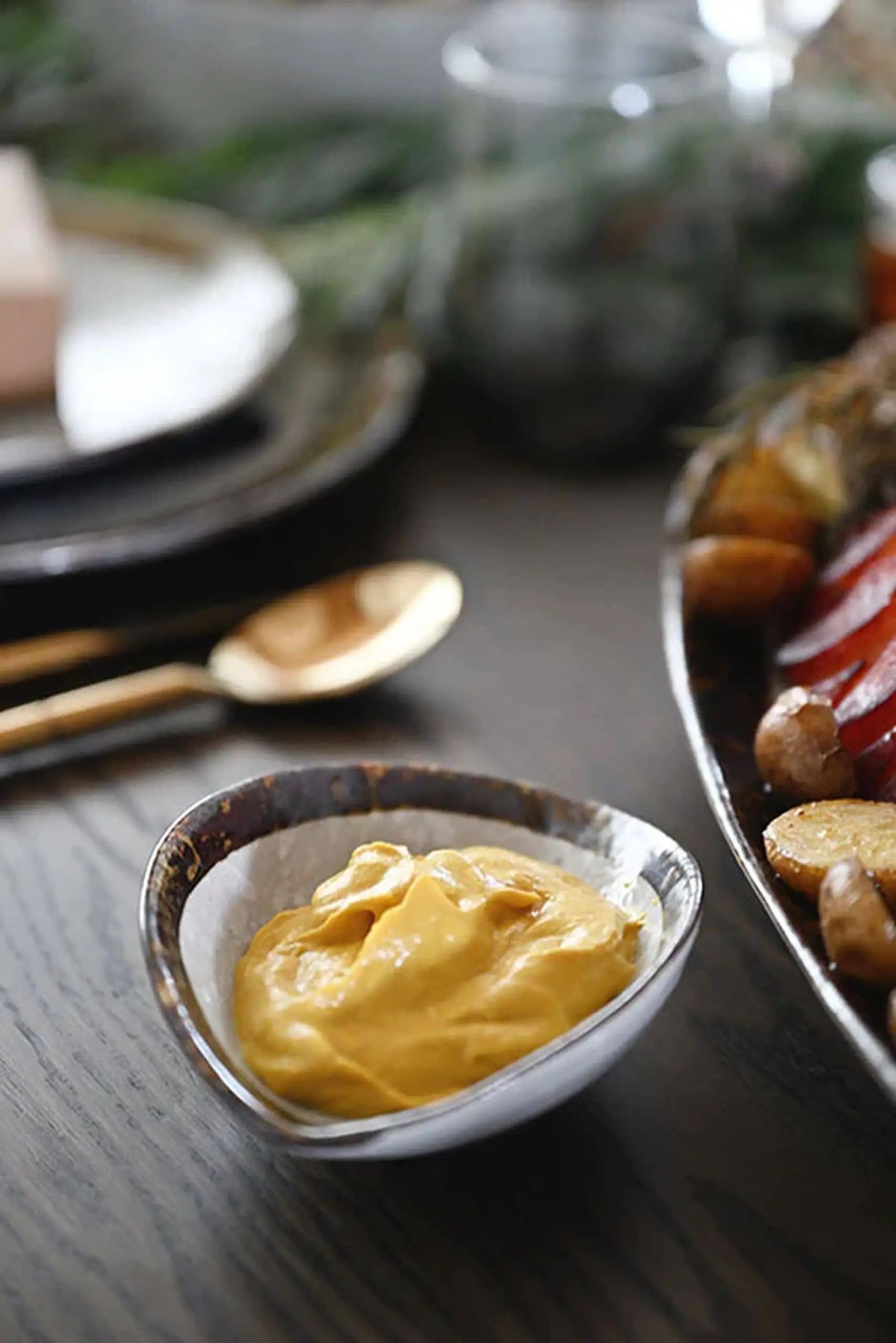 The Kra Sanctuary Satay Shell Dipping Dish by Kra Sanctuary holds creamy mustard on a dark wooden table, placed beside roasted potatoes and vegetables, with earthy dinnerware and a gold spoon gleaming in the background.