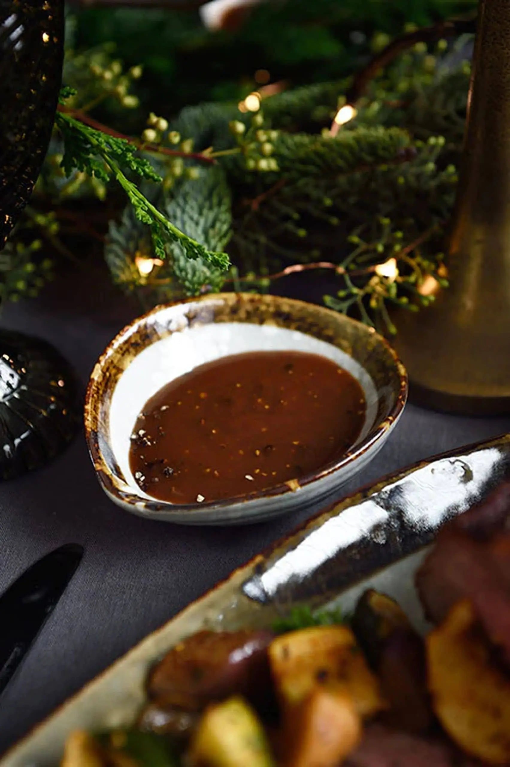 The Kra Sanctuary Satay Shell Dipping Dish by Kra Sanctuary, a rustic ceramic bowl with an iridescent border, holds brown gravy on a dark table amid festive greenery and string lights, with a nearby plate of food partially visible.