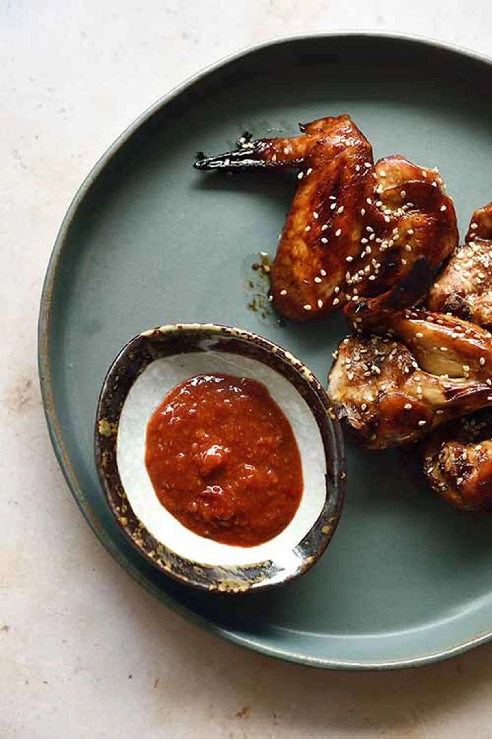 Glazed chicken wings with sesame seeds rest on earthy dinnerware, accompanied by a Kra Sanctuary Satay Shell Dipping Dish from Kra Sanctuary filled with reddish sauce.