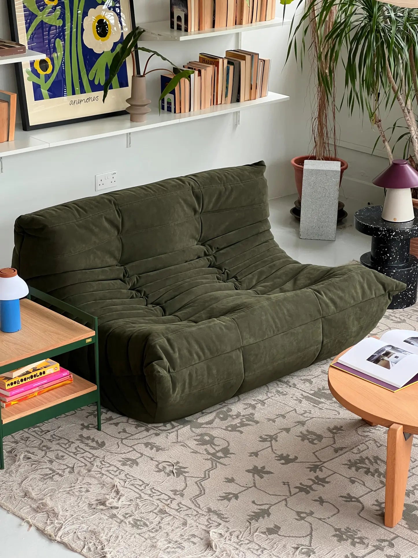 A cozy living room with a green Ligne Roset Togo 2 Seater loveseat, pleated fabric design, a light patterned rug, wooden coffee table with books, green side table, houseplants, and shelves displaying books and artwork.