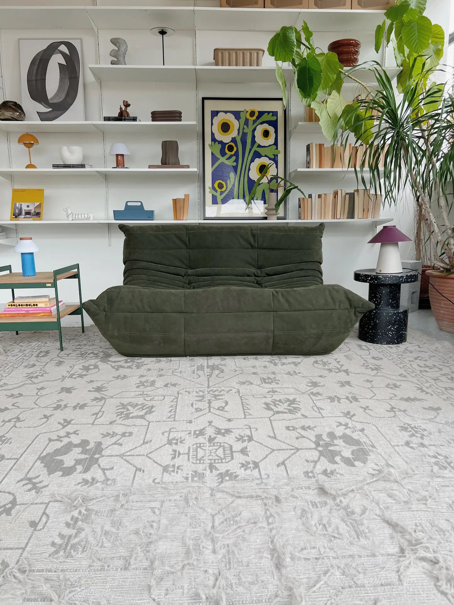 A modern living room with a Ligne Roset Togo, 2 Seater sofa by Ligne Roset featuring pleated upholstery, a patterned rug, potted plants, white shelving with books and decor, and vibrant floral wall art.