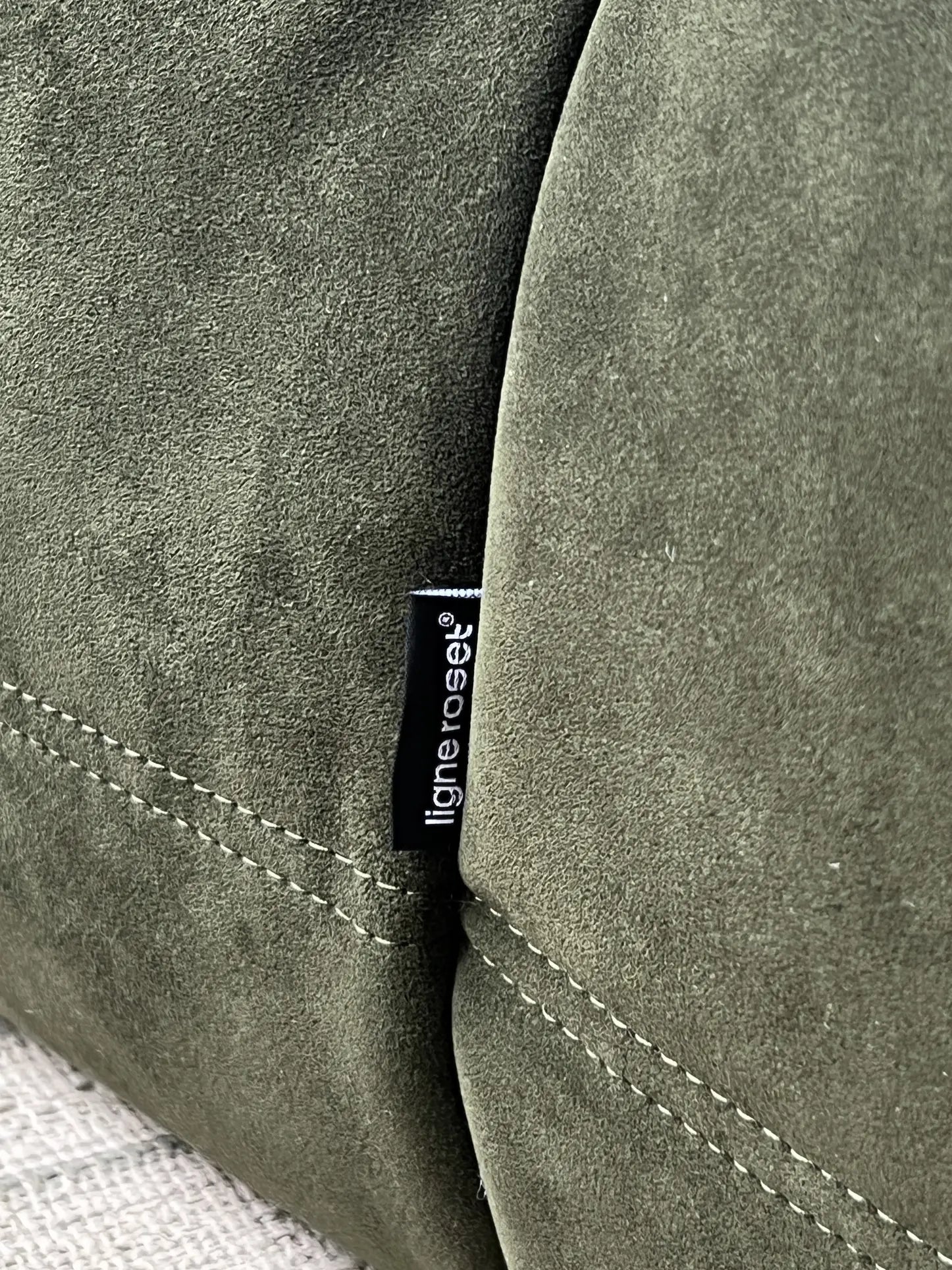 Close-up of olive green suede upholstery with visible stitching and a small black fabric tag reading Ligne Roset, highlighting the iconic pleated design of the Ligne Roset Togo, 2 Seater sofa.