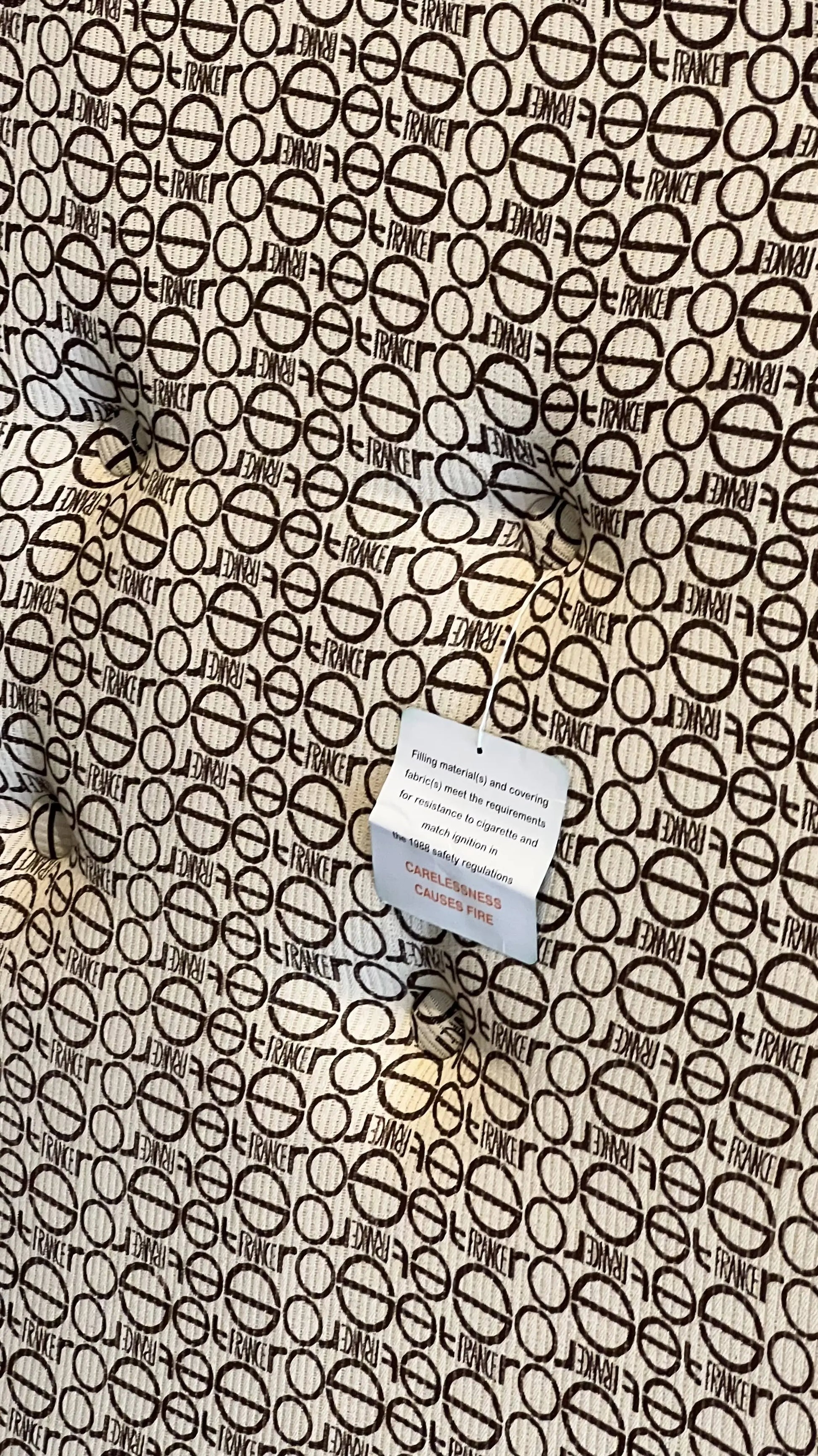 A close-up of the Ligne Roset Togo, 2 Seater by Ligne Roset shows a beige and brown mid-century geometric pattern with circles, lines, and FRANCE printed repeatedly. A white fabric tag with red and black text is attached to the piece.