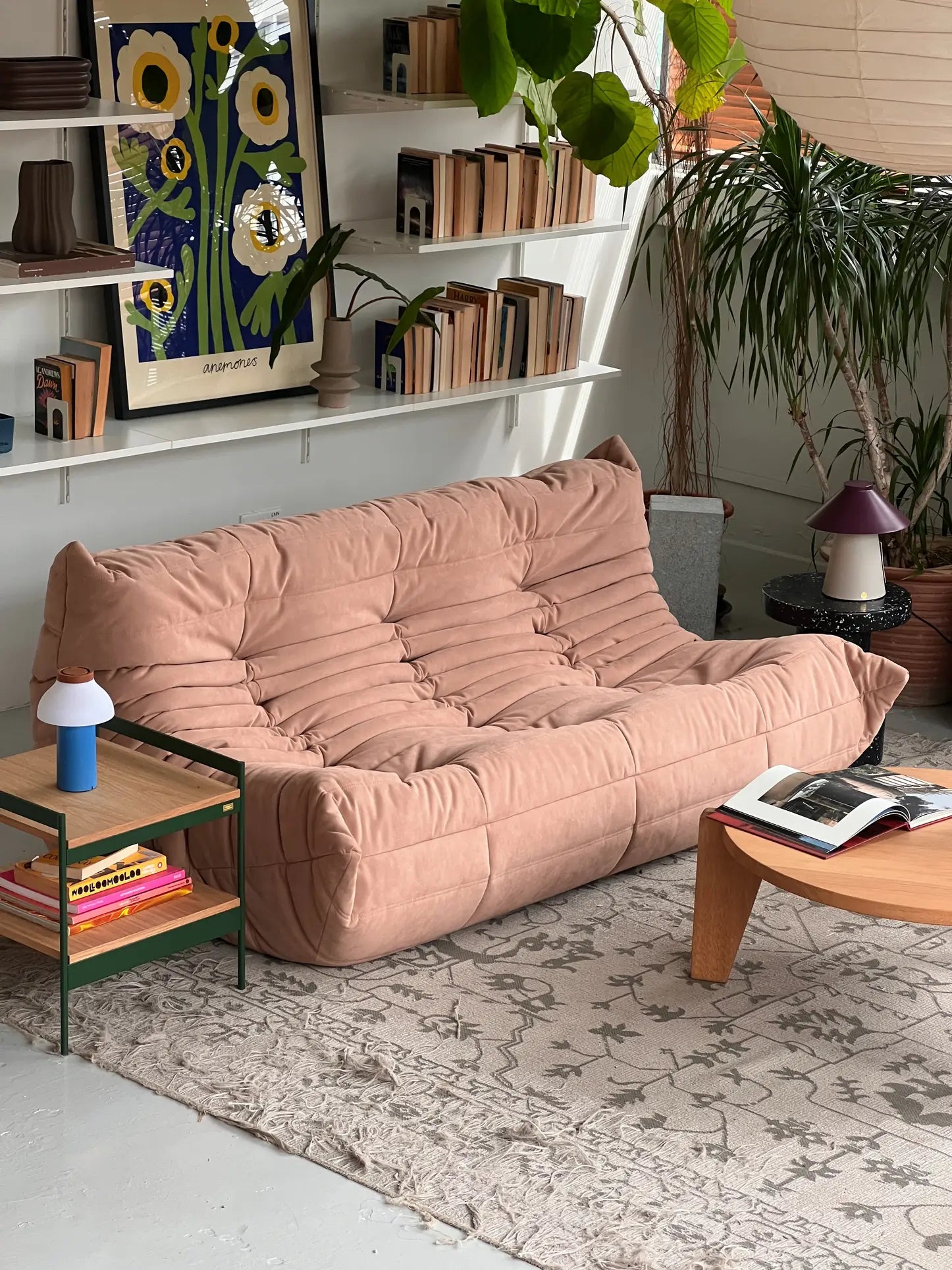 A cozy living room with a light pink Ligne Roset Togo 3 Seater sofa, pleated fabric, a wooden coffee table, geometric rug, green plants, shelves of books, colorful art, plus a side table displaying a blue lamp and stacked magazines.