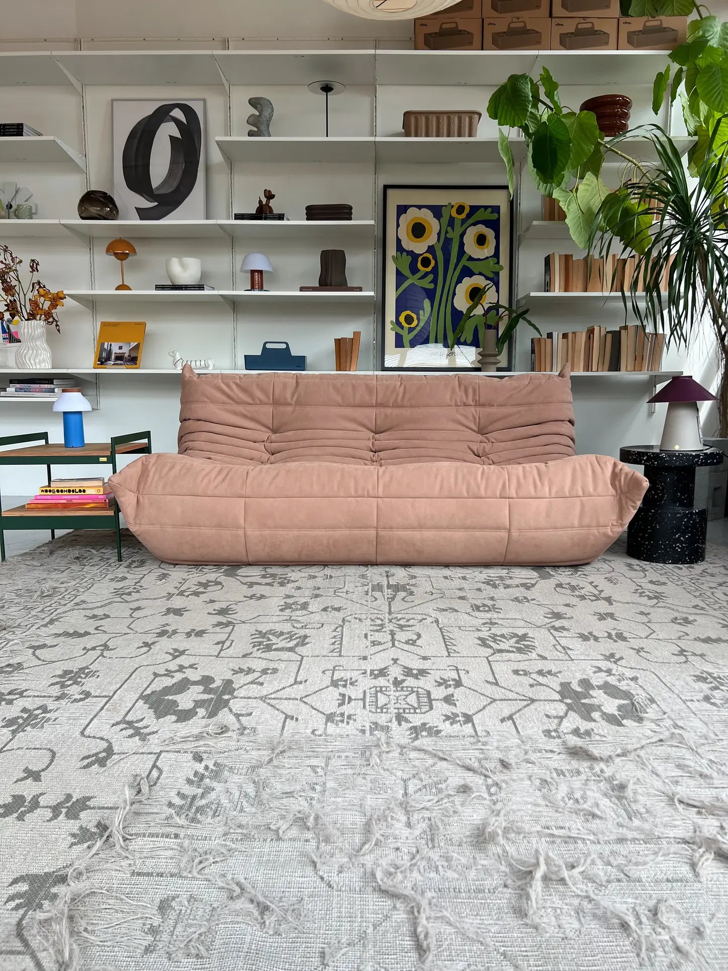 A modern living room with a Ligne Roset Togo, 3 Seater sofa in pink, patterned gray rug, white book-filled shelves, abstract art, plants, and colorful table lampsโall blending mid-century accents.