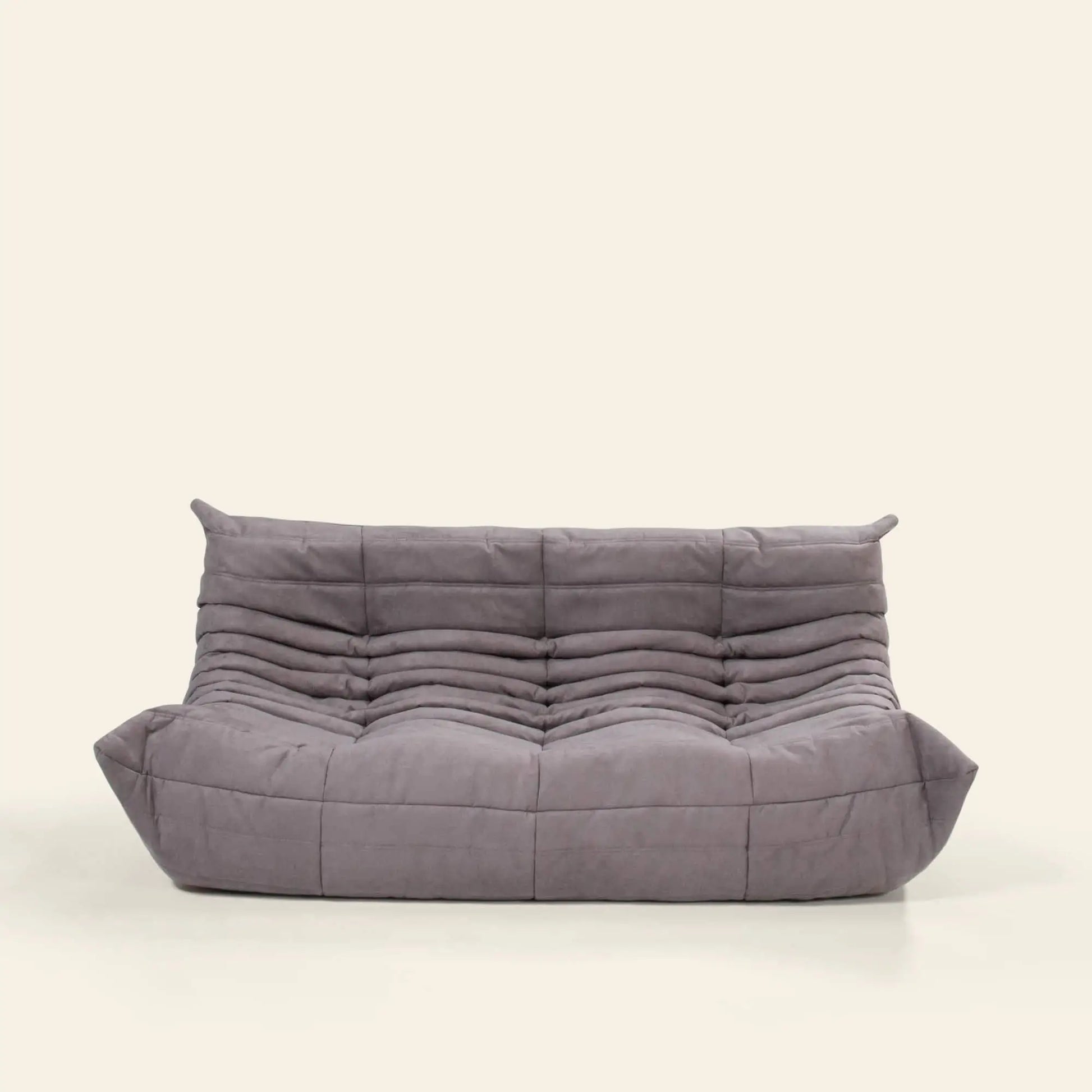 The Ligne Roset Togo, 3 Seater by Ligne Roset features a modern low-profile gray design with plush tufting and wide pillow-like armrests and backrest, bringing mid-century flair to a simple light backdrop.