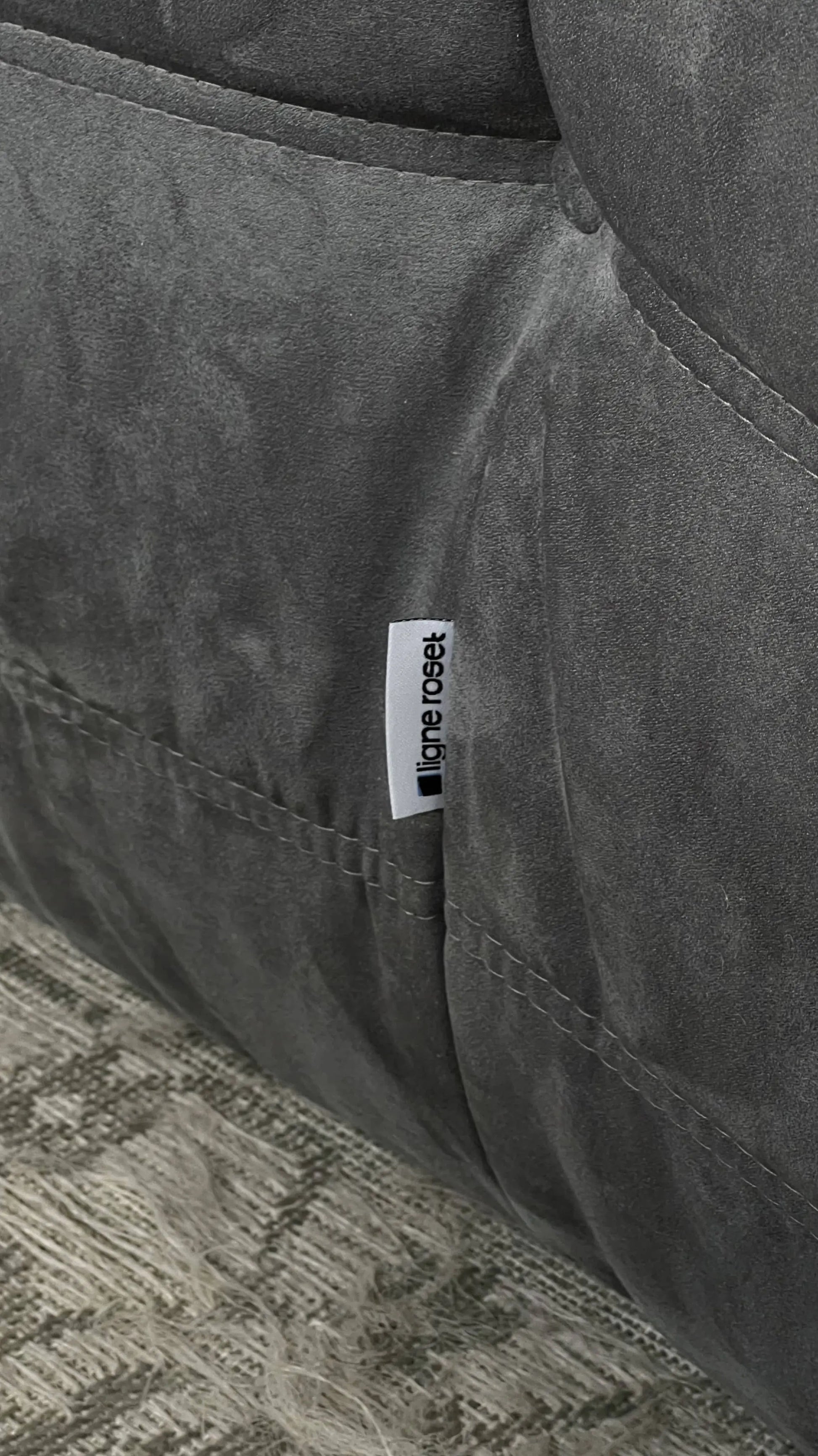 A close-up of the Ligne Roset Togo, 3 Seater by Ligne Roset, showcasing its modular gray suede design with a fabric tag stitched into the seam as it sits on a textured gray carpet.