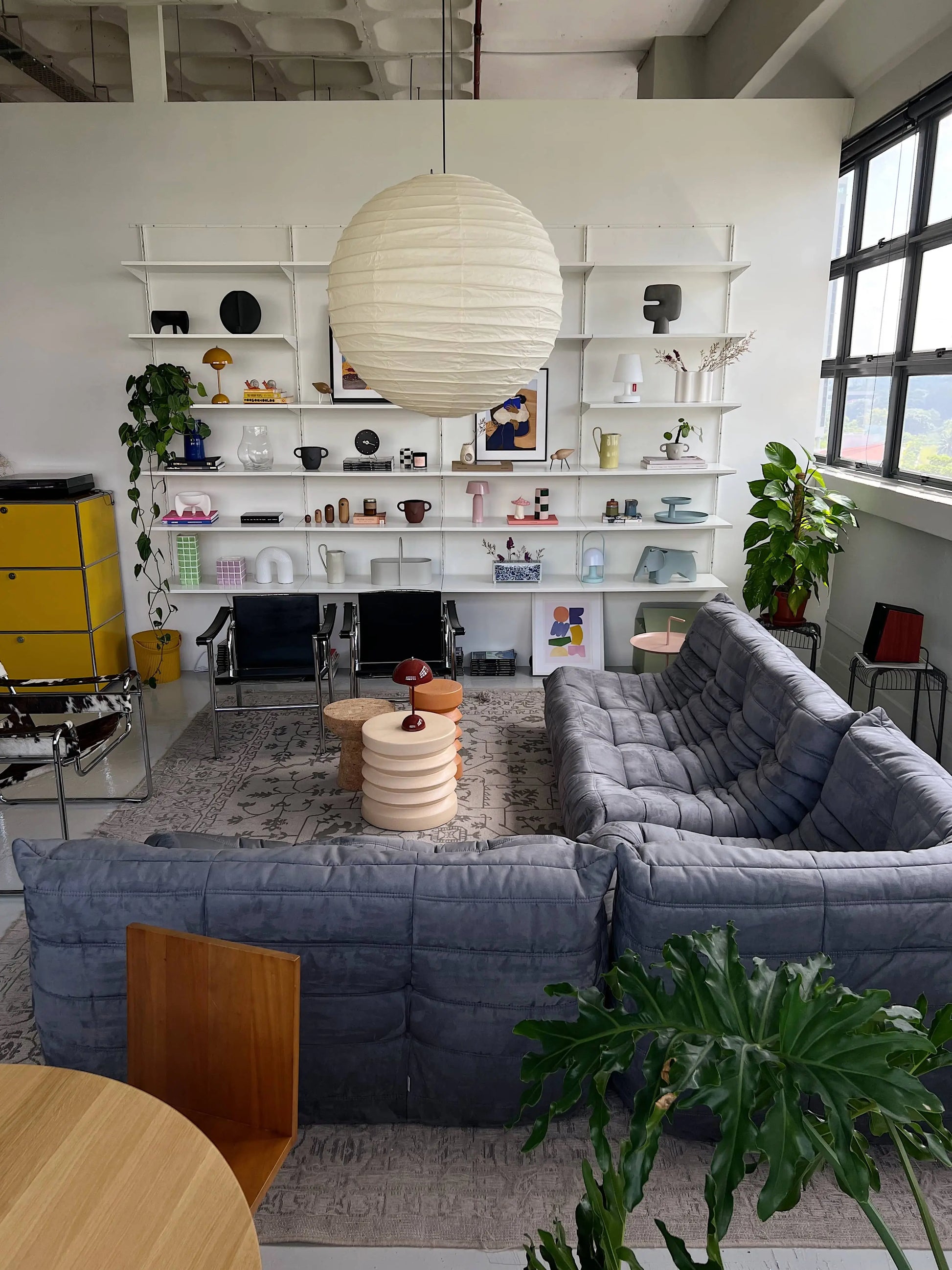 A modern living room showcases the Ligne Roset Togo, 3 Seater by Ligne Roset in gray, a round paper lantern ceiling light, open shelves with decor, plants, a yellow cabinet, and large windows—embracing mid-century style with vibrant accents.