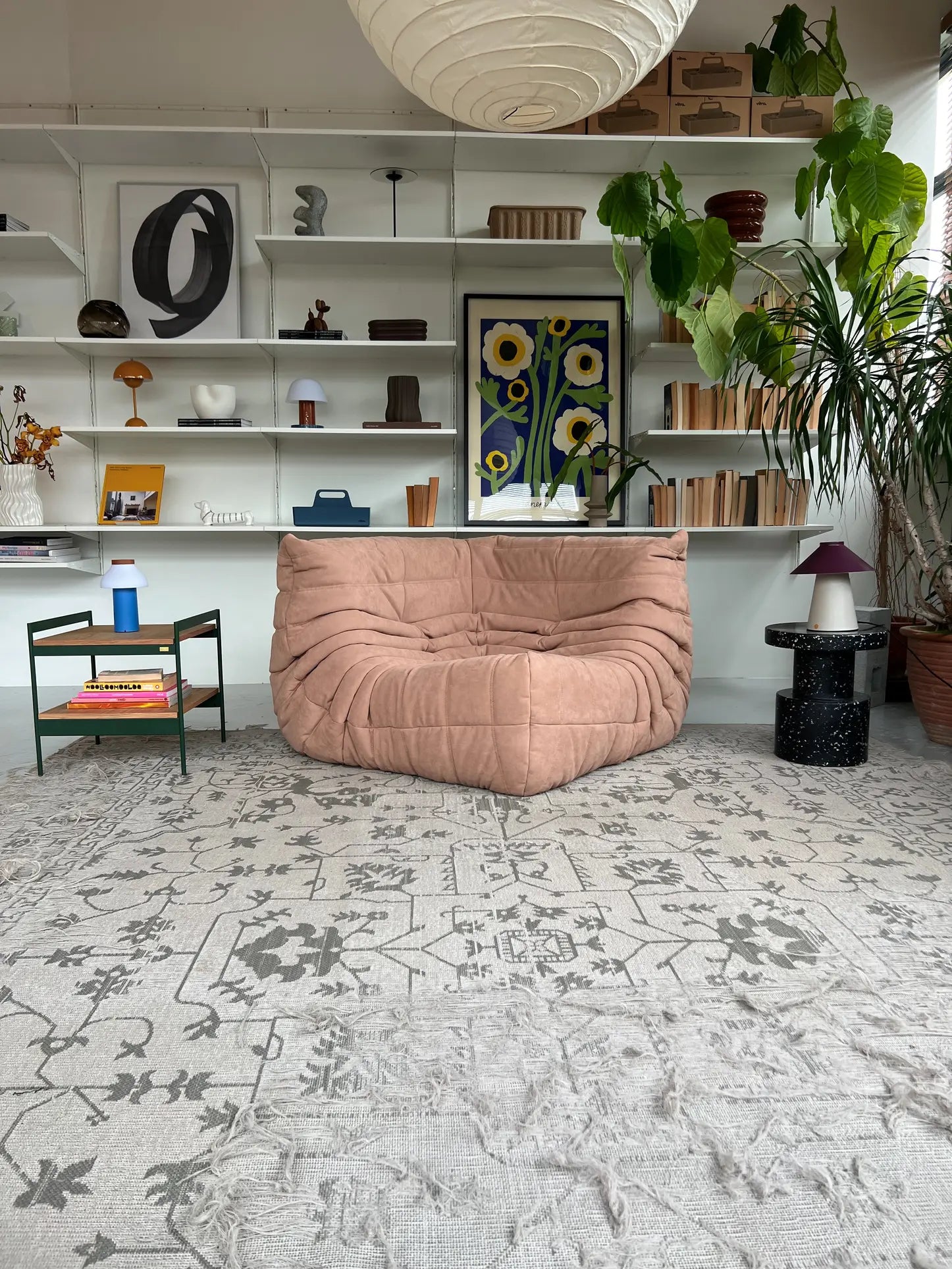 A cozy living room features a tan Ligne Roset Togo Corner Seat, mid-century style, on a patterned gray rug. A green side table stands beside bookshelves with decor, books, and plants. A floral art print and large potted plant sit in the background.