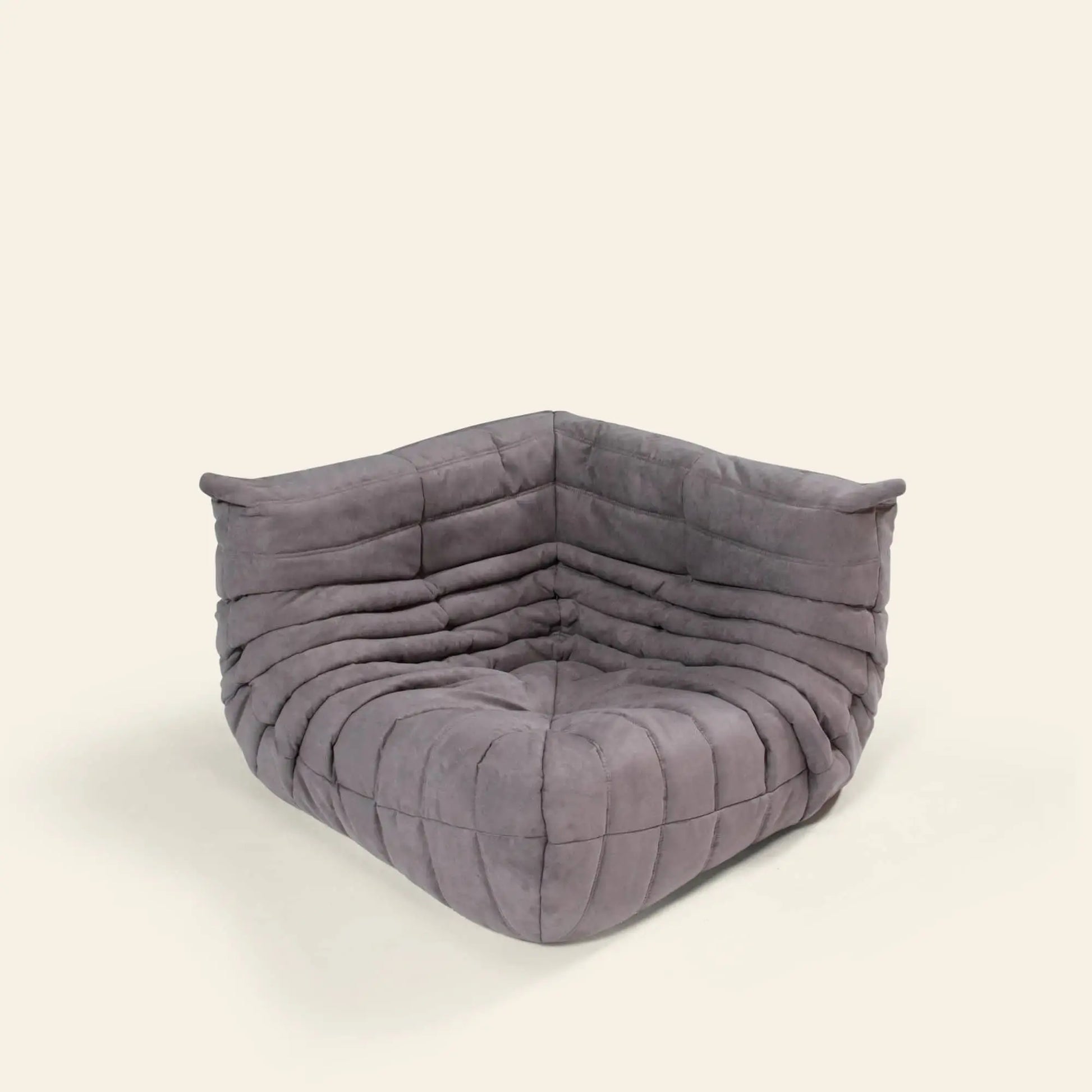 The Ligne Roset Togo Corner Seat by Ligne Roset is a modern, armless piece inspired by mid-century design, upholstered in soft gray fabric with horizontal and vertical stitching, shown against a plain light background.