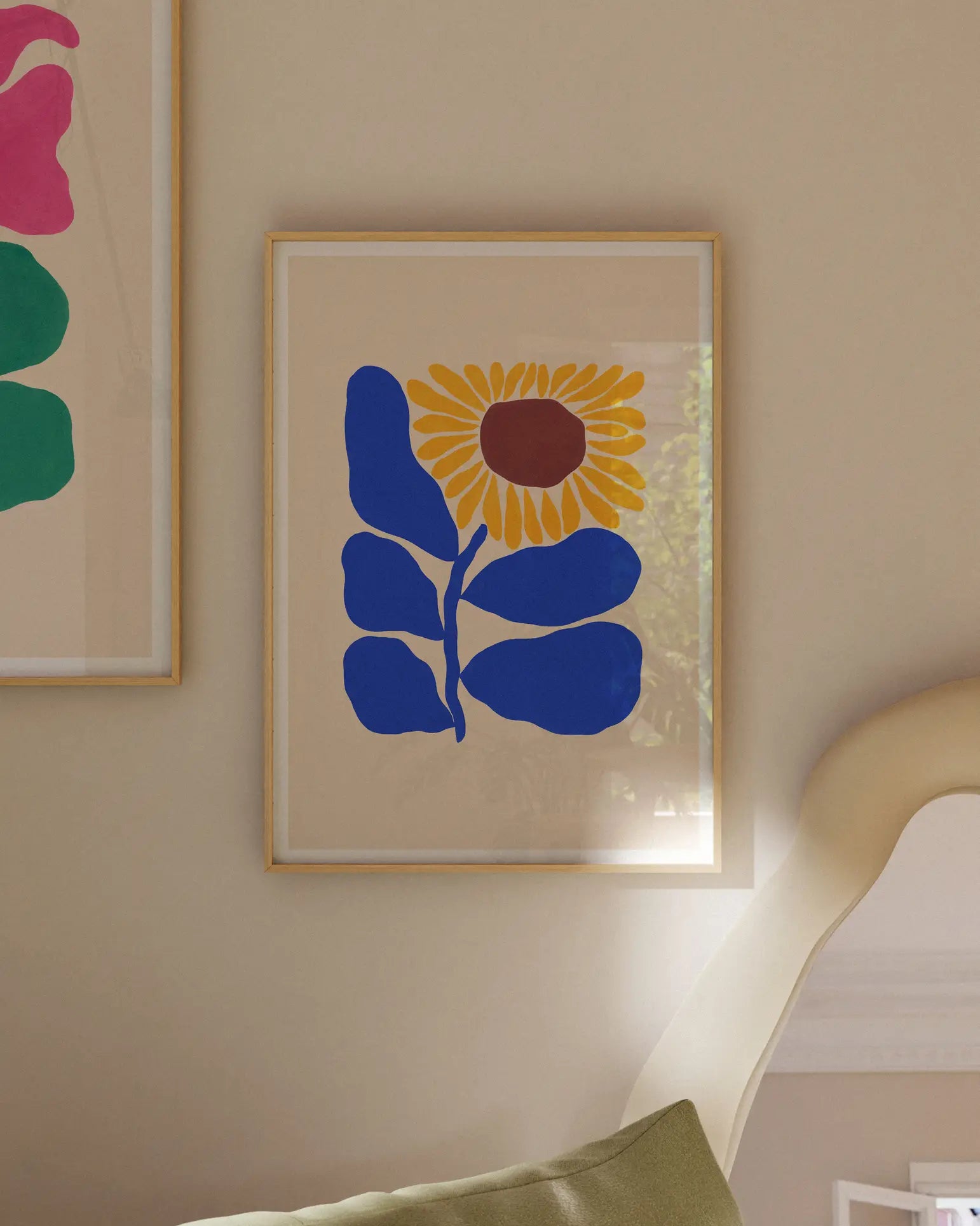 The Liv Lee Sunflower, an A3 framed archival-quality giclée print on Hahnemühle stock by Liv Lee, features a bold abstract sunflower and hangs above a green pillow and curved chair. Another colorful artwork is partially visible on the left.