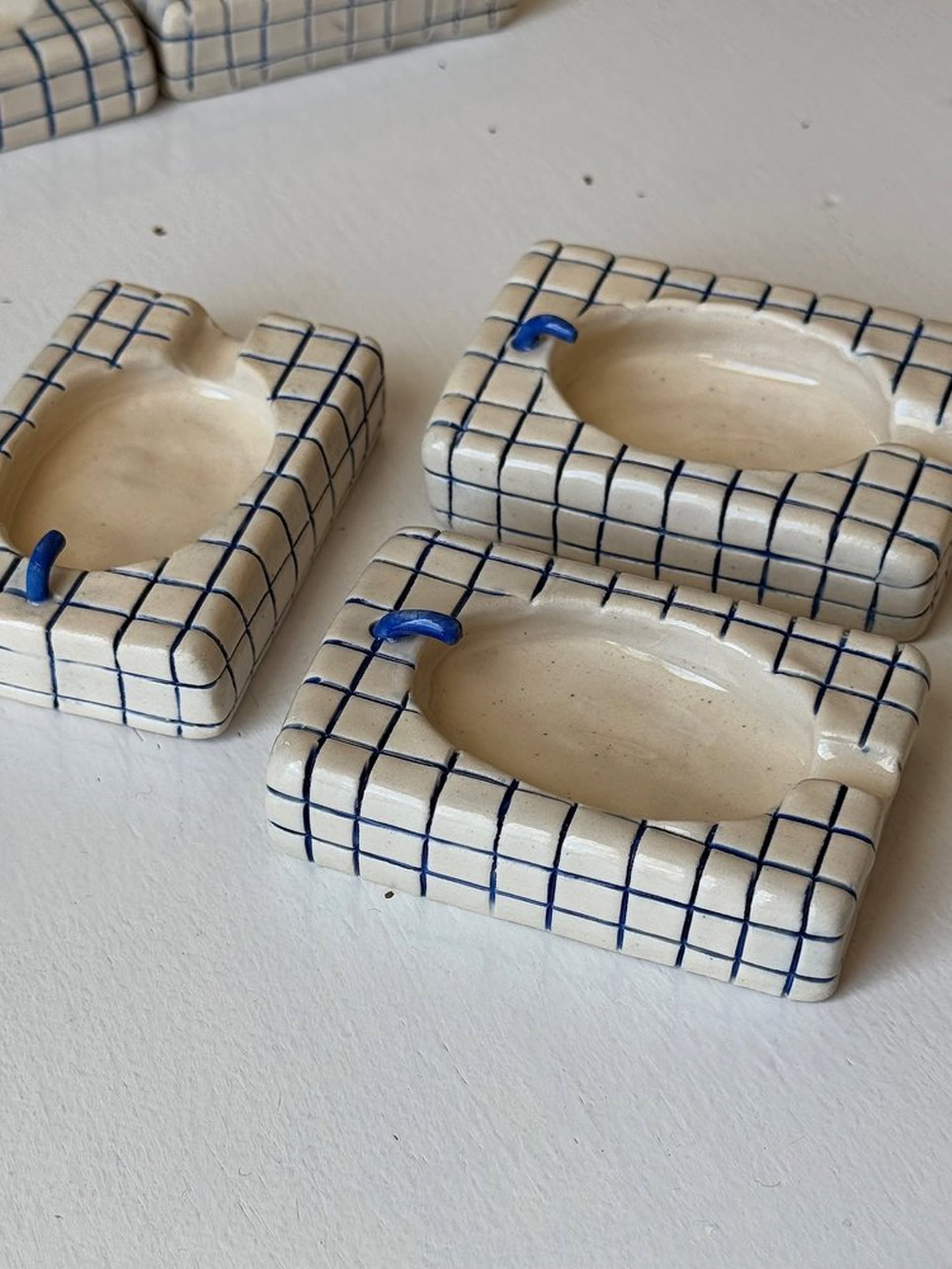 The MIRYO Bathtub Ashtray by MIRYO features a white base with blue grid patterns and a blue accent notch for cigarettes, evoking retro motel vibes. Three handmade rectangular ashtrays are displayed on a white surface.
