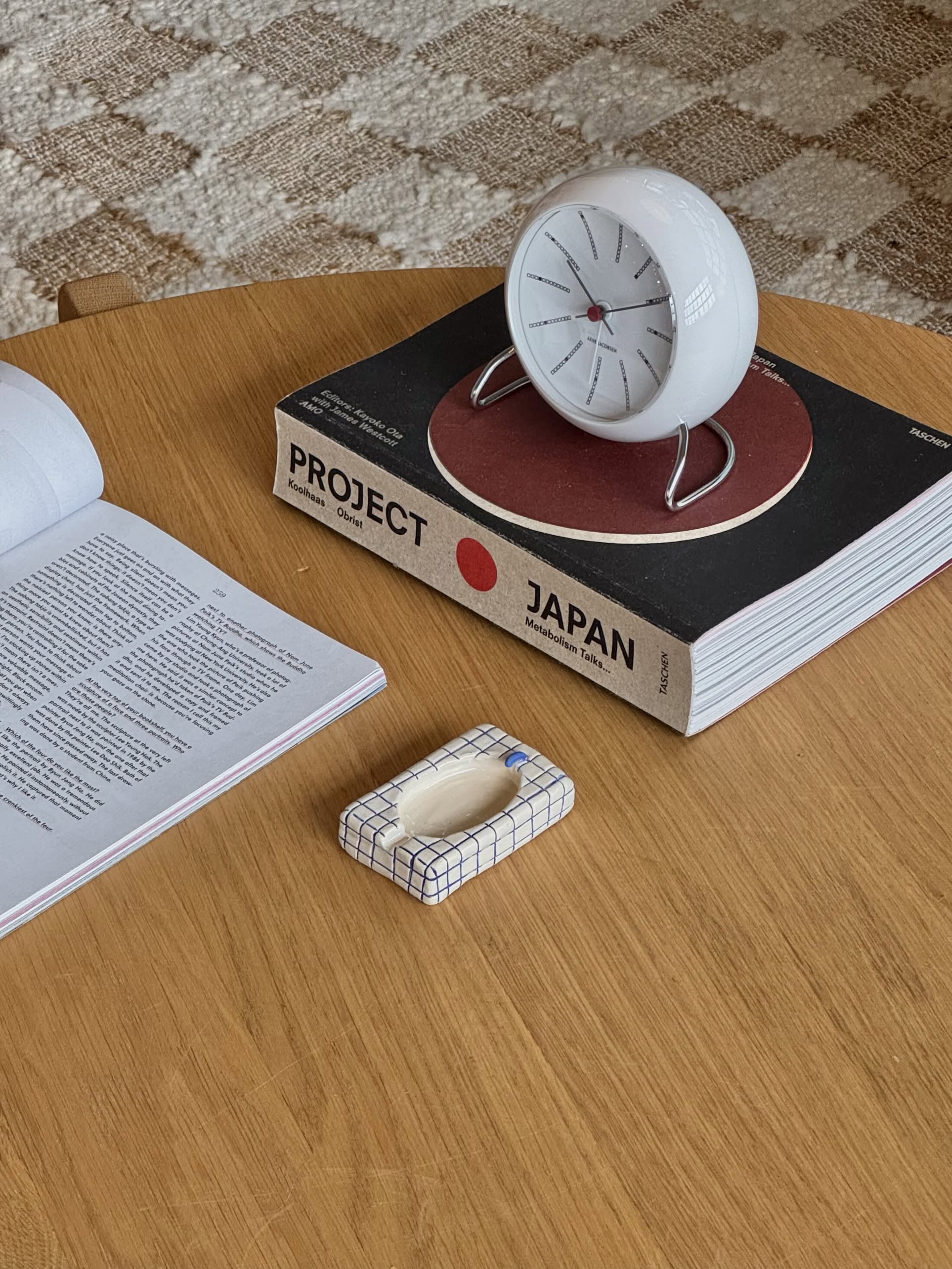 A round white clock, an open book, and the MIRYO Bathtub Ashtray by MIRYO are on a wooden table. The clock is placed on a large book titled Project Japan. A light checkerboard carpet lies underneath.