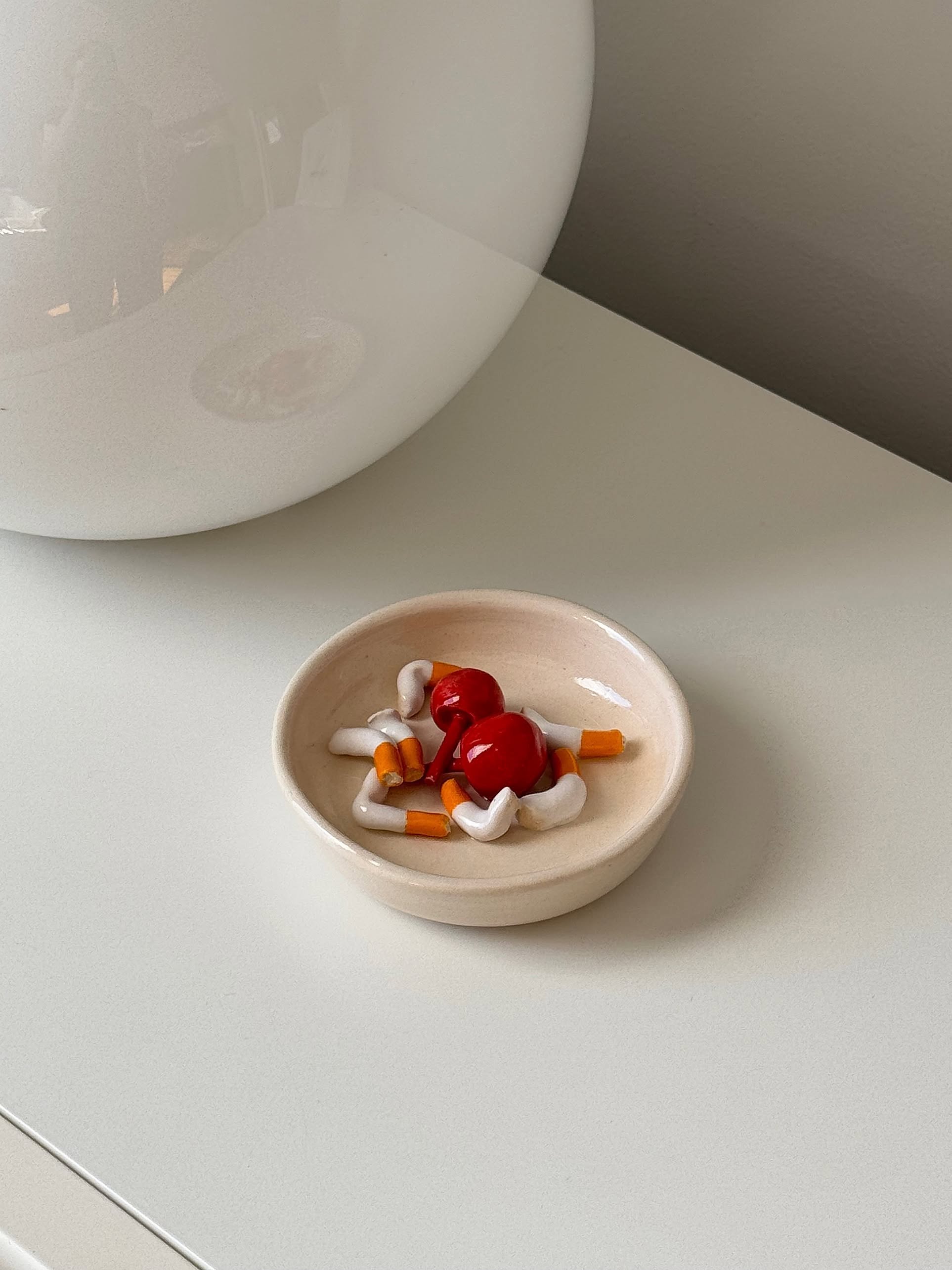 A small beige MIRYO Miss Cherries and Cigs Ashtray holds colorful, toy-like mushroom accents, adding a unique decorative touch to a white surface near a round, reflective lamp.