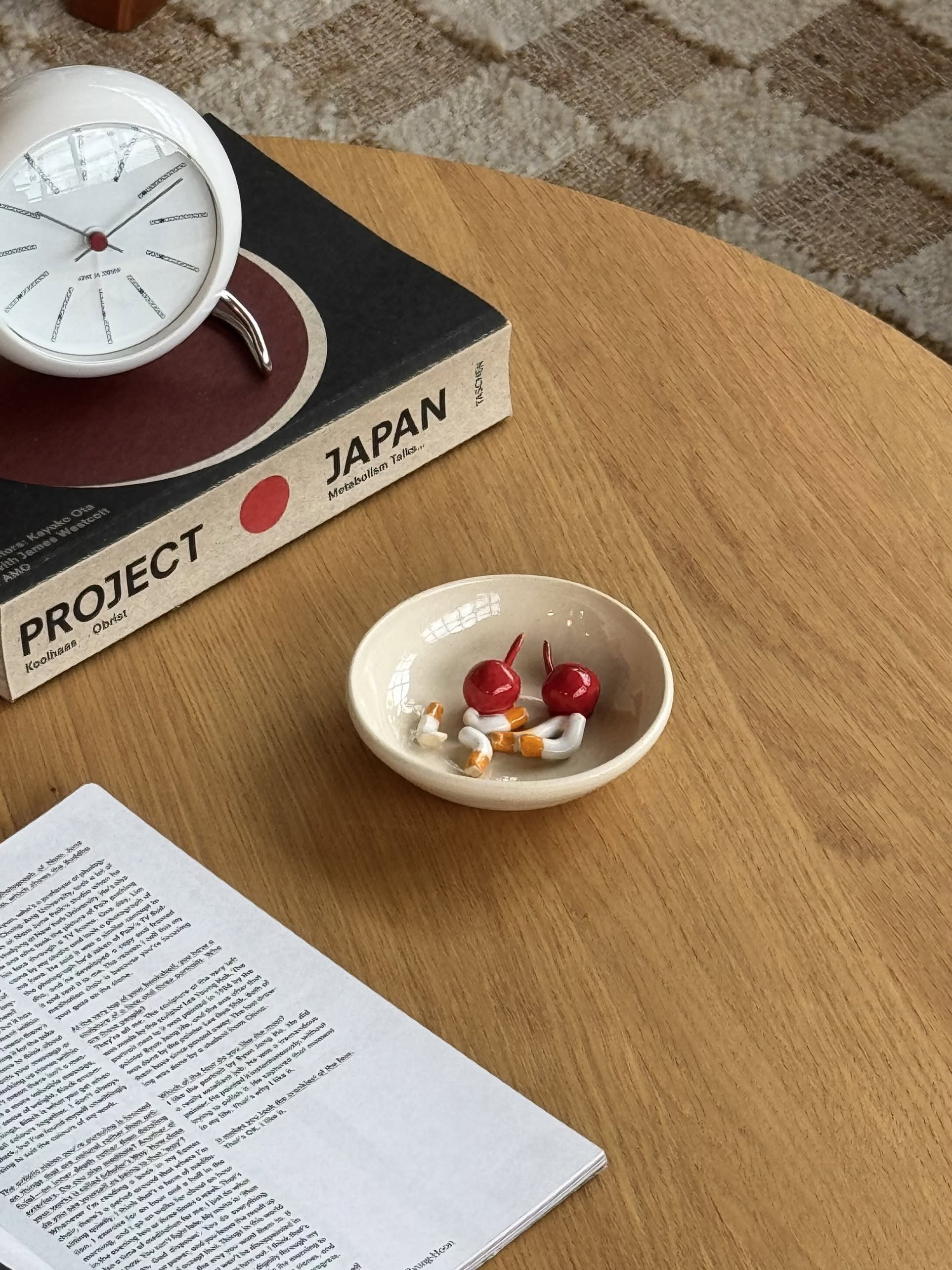 A unique decorative accent, this small bowl with apple and garlic miniatures sits on a wooden table near an open book, a clock, and Project Japan—a charming touch reminiscent of the MIRYO Miss Cherries and Cigs Ashtray.
