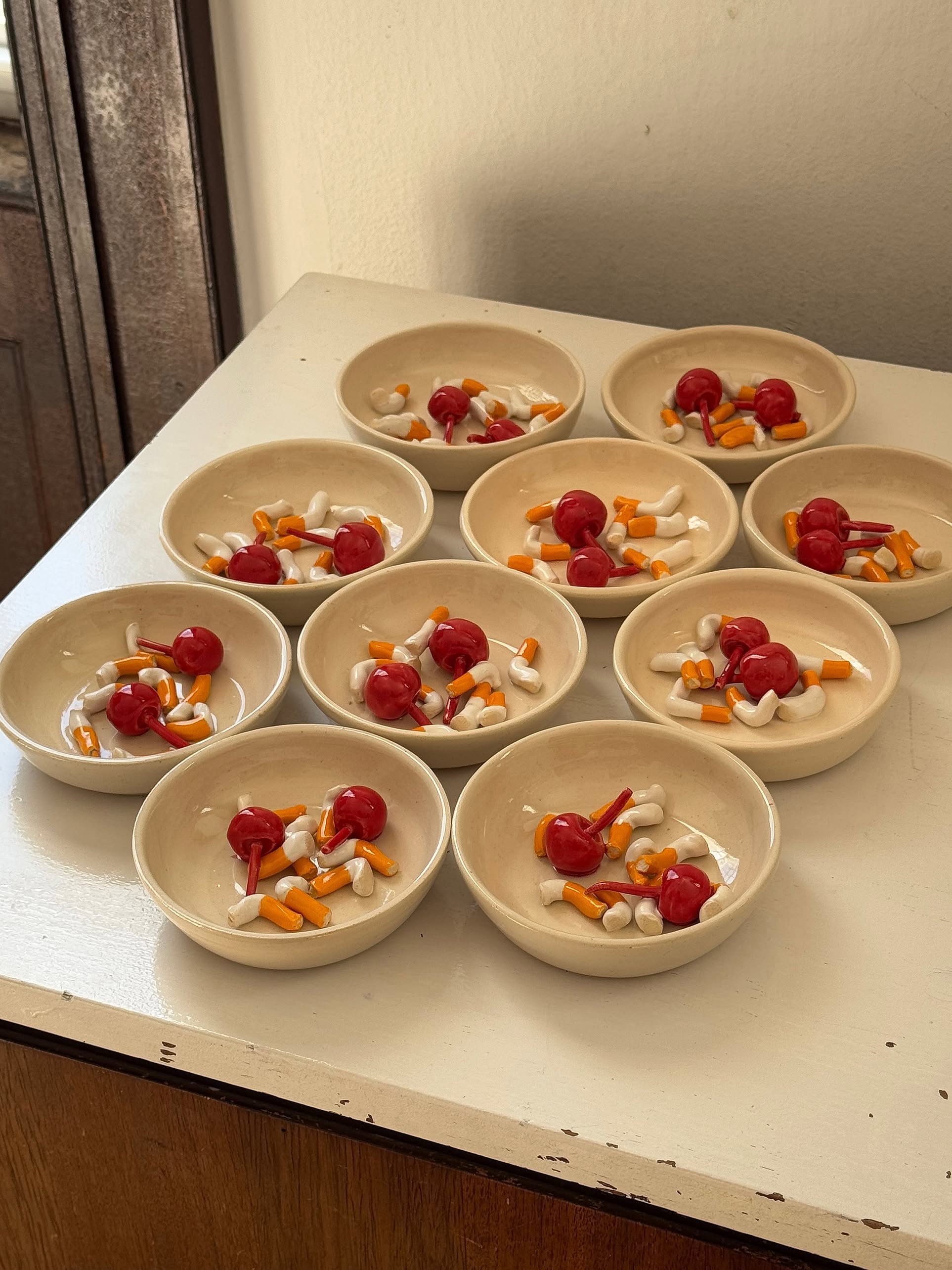 Ten cream-colored MIRYO Miss Cherries and Cigs Ashtrays by MIRYO are displayed on a white surface, each filled with red, white, and orange pill-shaped accents—a quirky, decorative touch for any space.