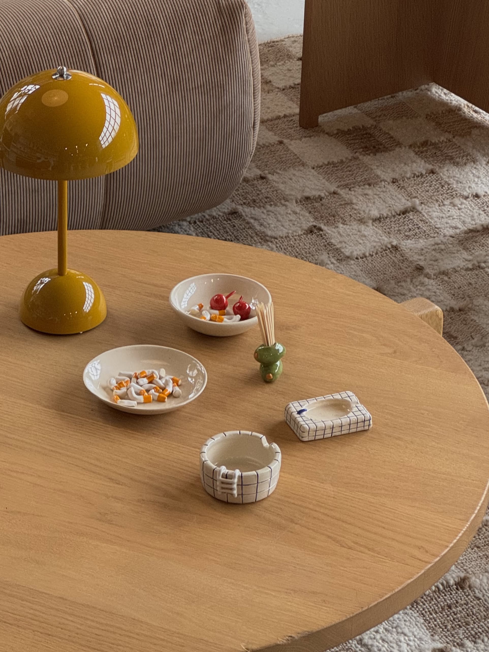 A round wooden coffee table features a yellow lamp, ceramic dishes with pills and cherry tomatoes, toothpicks in a holder, and the handcrafted MIRYO Miss Cherries and Cigs Ashtray by MIRYO. A patterned rug and a ribbed sofa are seen in the background.