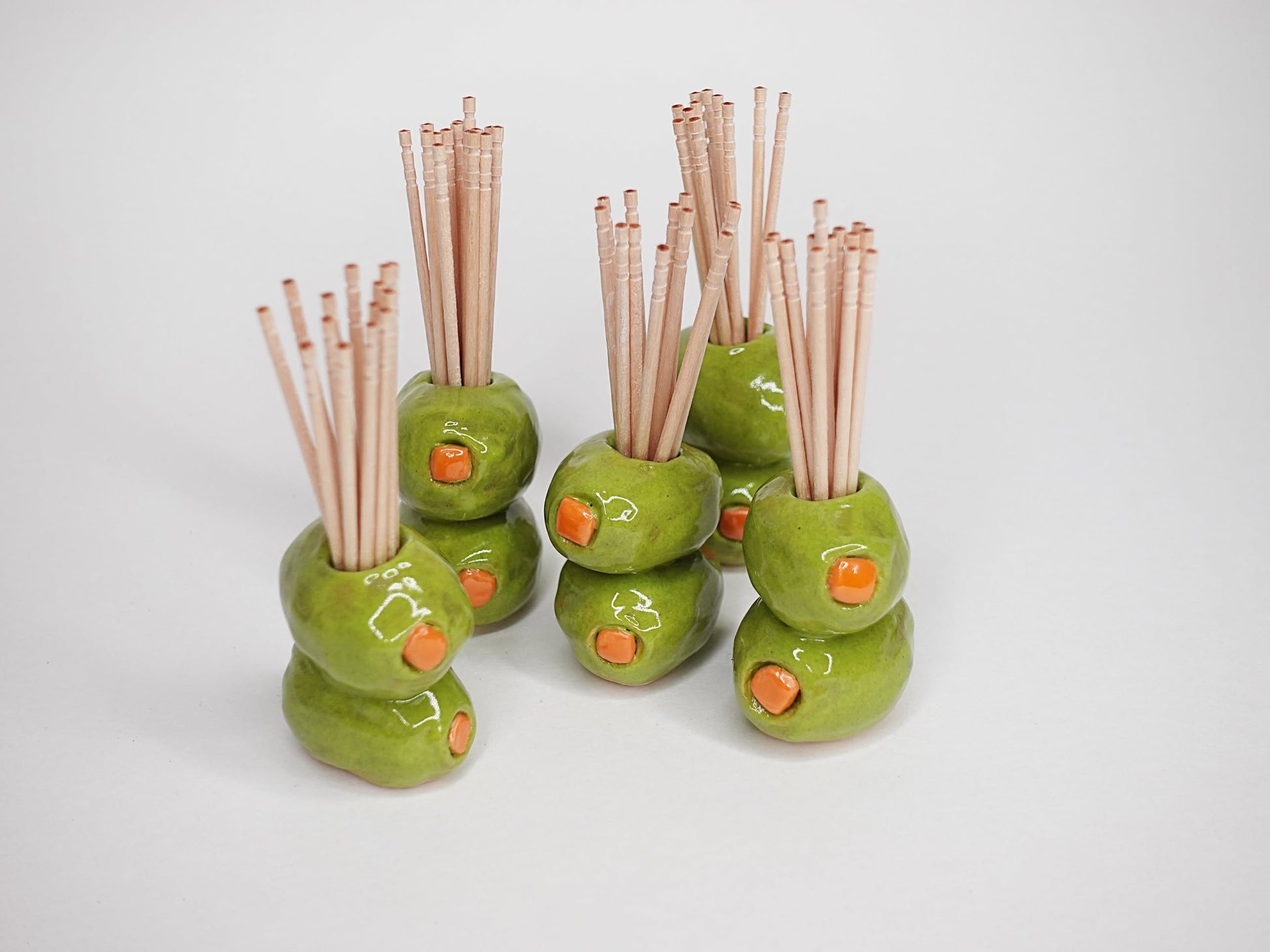 Several sets of wooden chopsticks stand upright in MIRYO Olive Toothpick Holders—ceramic holders by MIRYO shaped and painted like green olives with pimento centers—arranged on a plain white background.