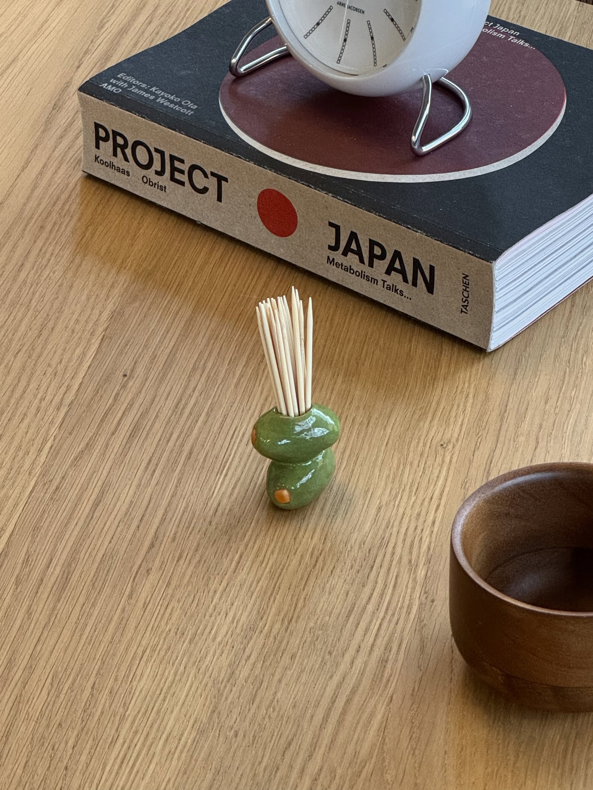 The MIRYO Olive Toothpick Holder by MIRYO, filled with toothpicks, a wooden bowl, and the large book Project Japan sit on a wooden table. A white clock rests atop the book.