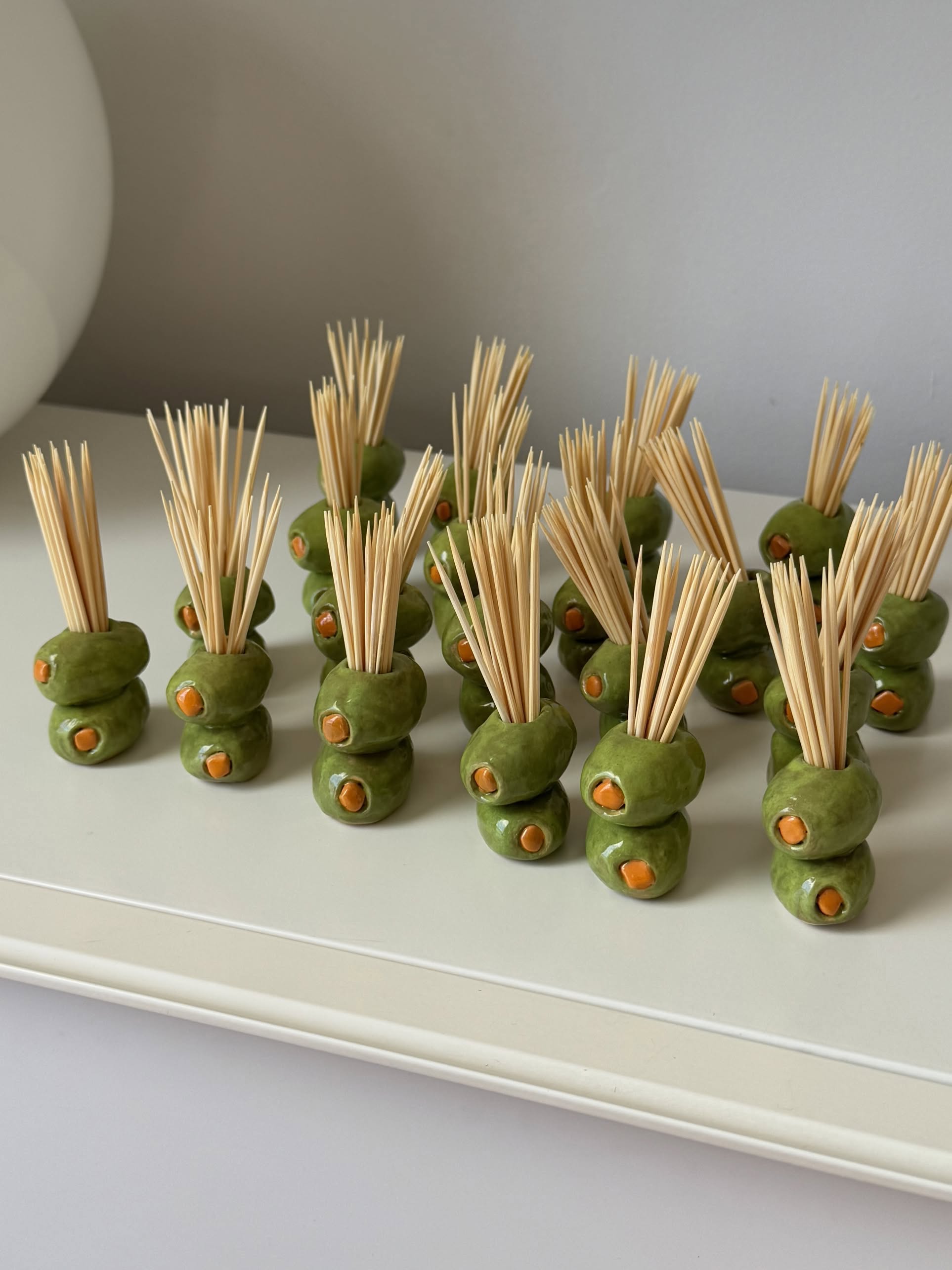 Several MIRYO Olive Toothpick Holders, handcrafted to resemble green olives with pimentos and designed to hold toothpicks upright, are arranged on a white tray against a neutral background.