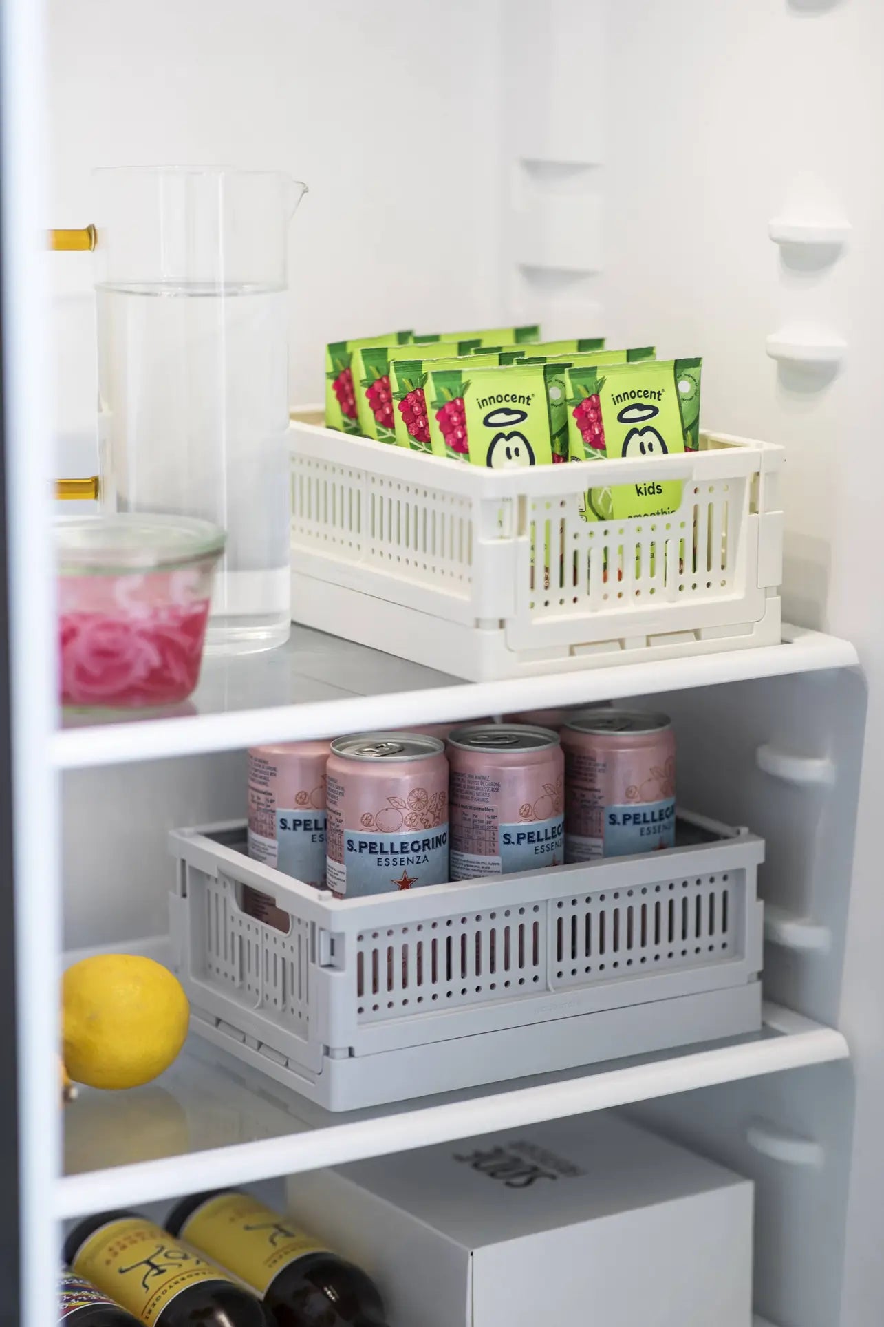 A neatly organized fridge shelf features juice pouches in a Made Crate Midi by Made Crate, a glass pitcher of water, a glass jar, canned San Pellegrino drinks in a gray recycled plastic crate, a lemon, and bottles on the lower shelf.