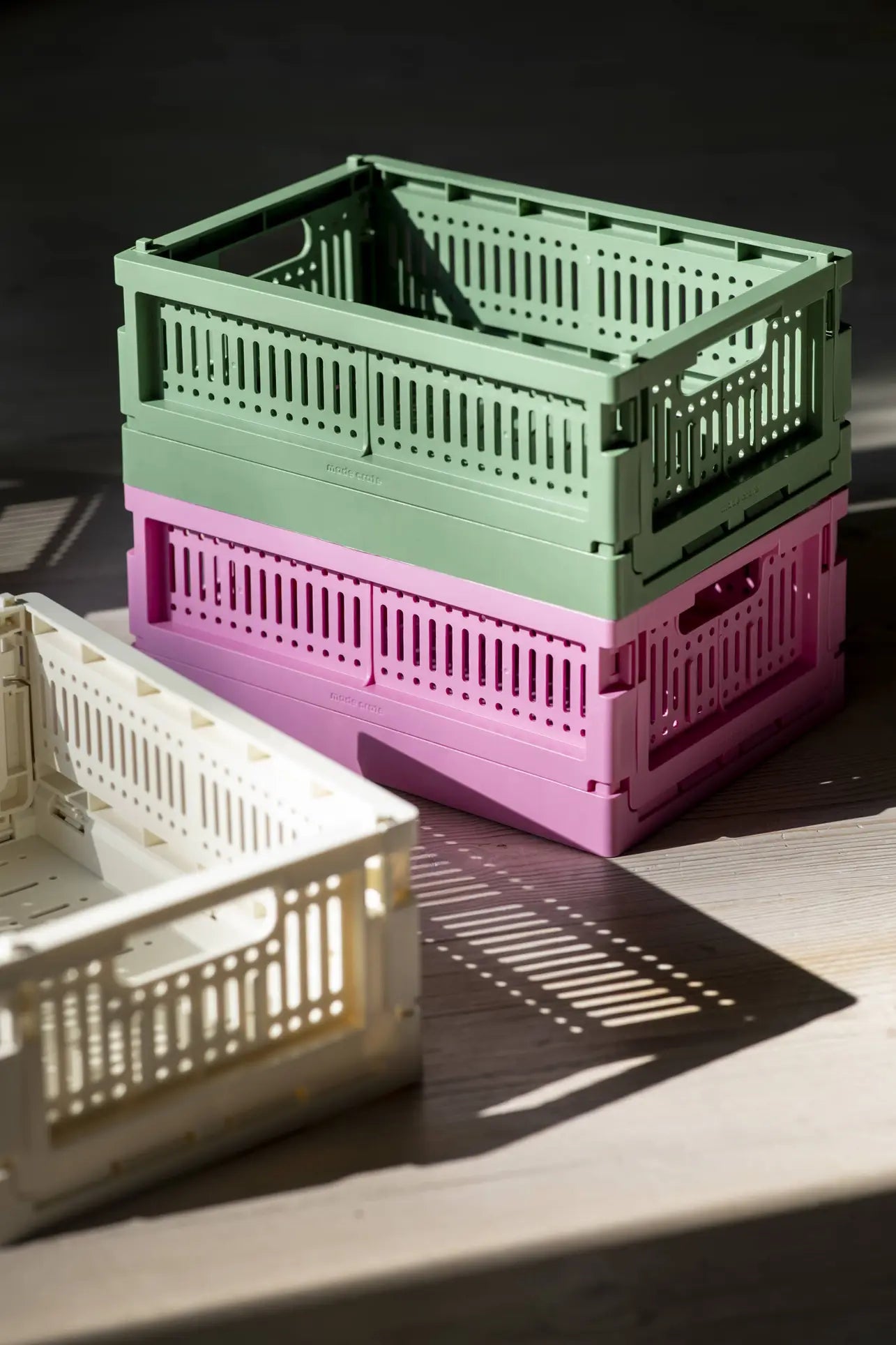 Three Made Crate Midi folding crates by Made Crate—green, pink, and off-white—are stacked on a sunlit wooden floor. Made from recycled plastic, they offer practical, eco-friendly storage and showcase convenient stacking.
