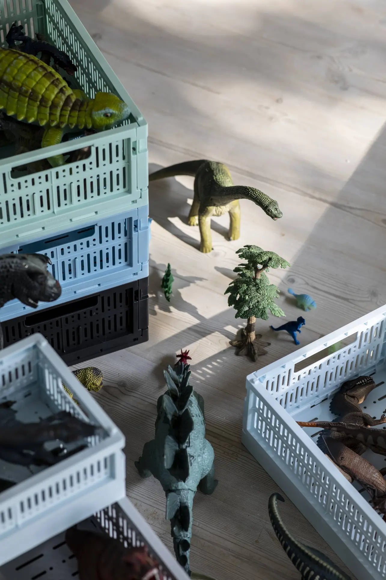 Toy dinosaurs and trees are displayed on a wooden floor beside Made Crate Midi eco-friendly folding crates by Made Crate, crafted from recycled plastic, with sunlight casting shadows across the scene.
