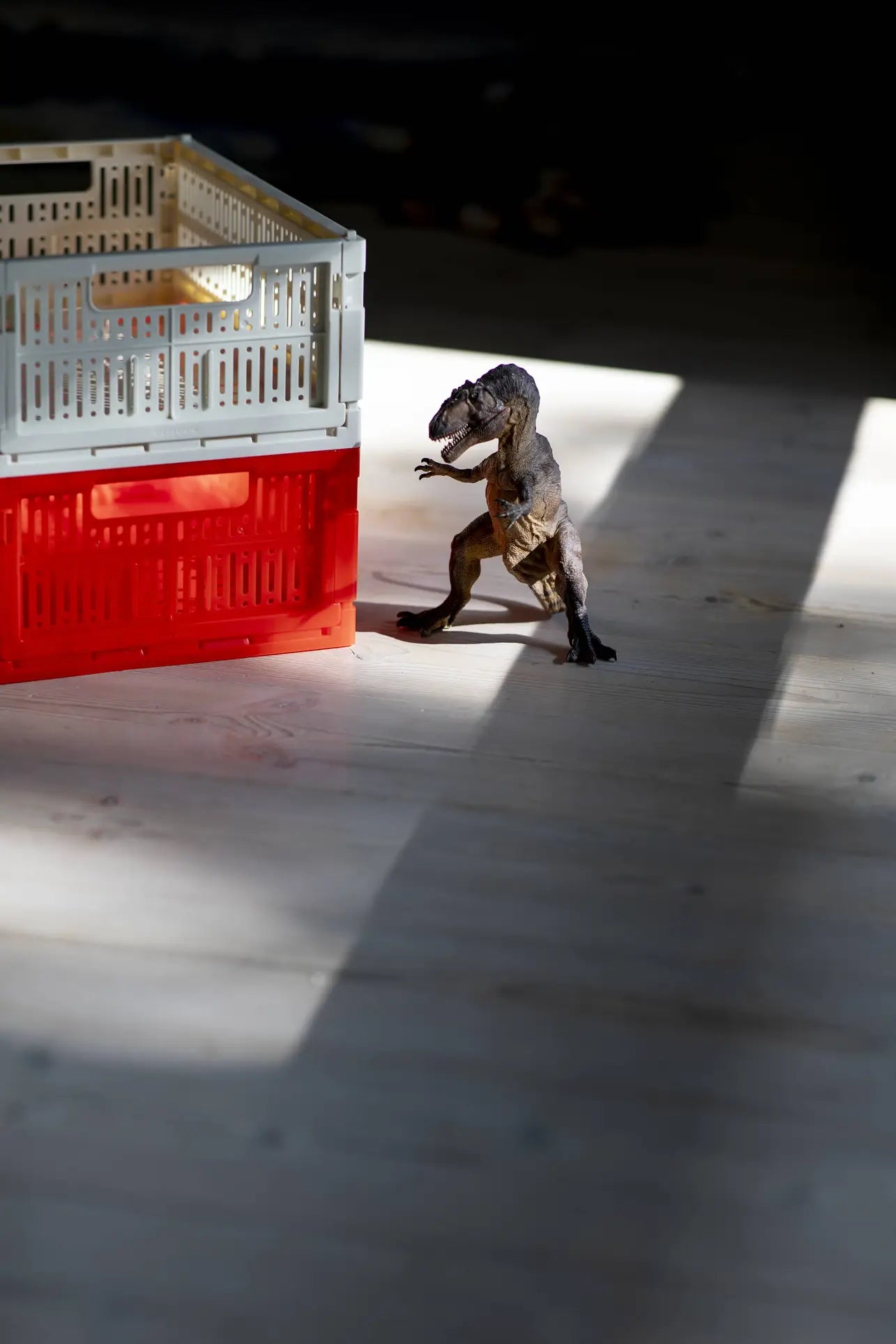 A small toy dinosaur stands upright on a wooden floor beside a Made Crate Midi eco-friendly folding crate by Made Crate, with sunlight streaming in and casting shadows across the scene.