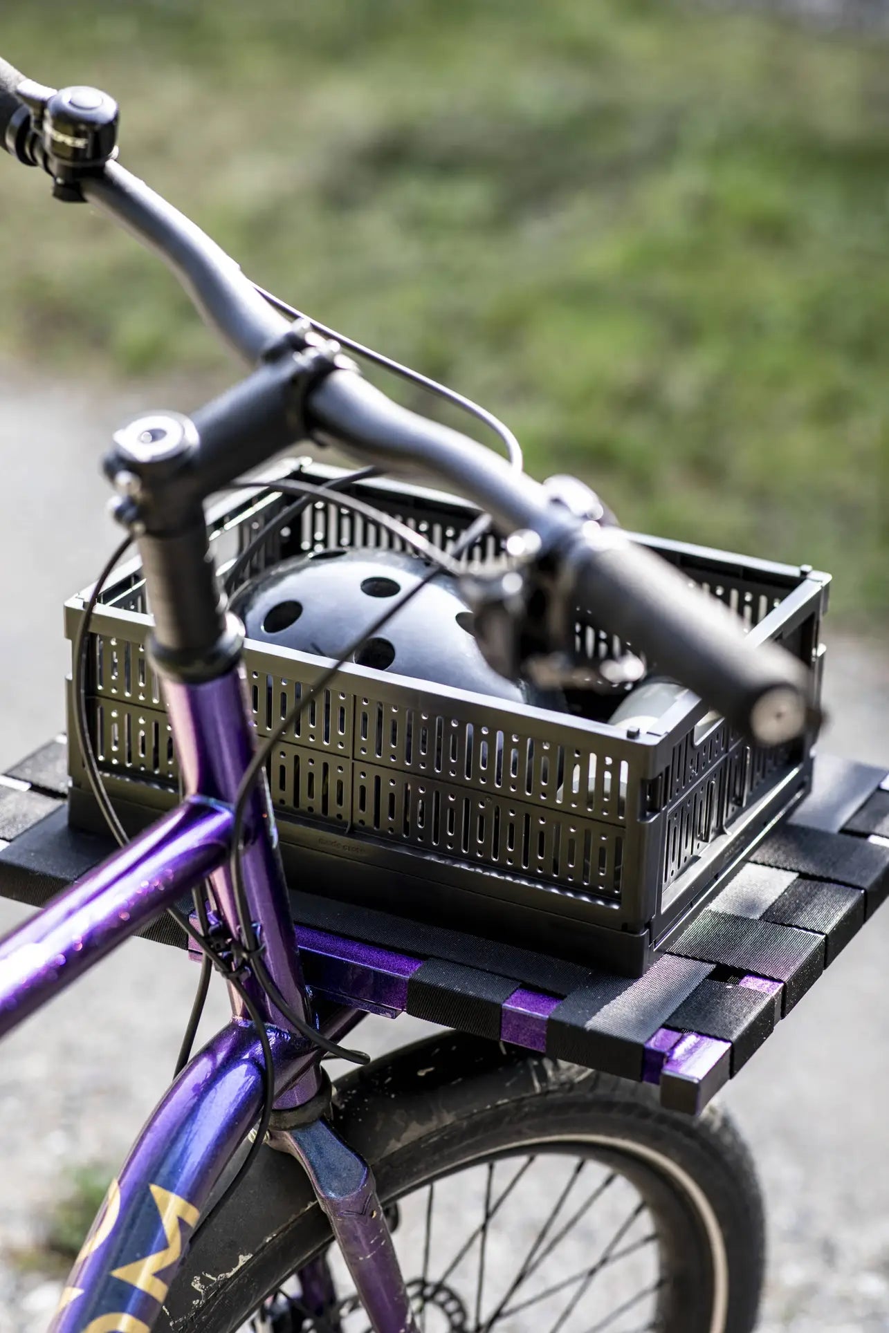 A close-up of a purple bicycle with a Made Crate Midi by Made Crate, an eco-friendly folding crate, attached to the front rack and holding a black helmet. The bike is on an outdoor path with grass in the background.