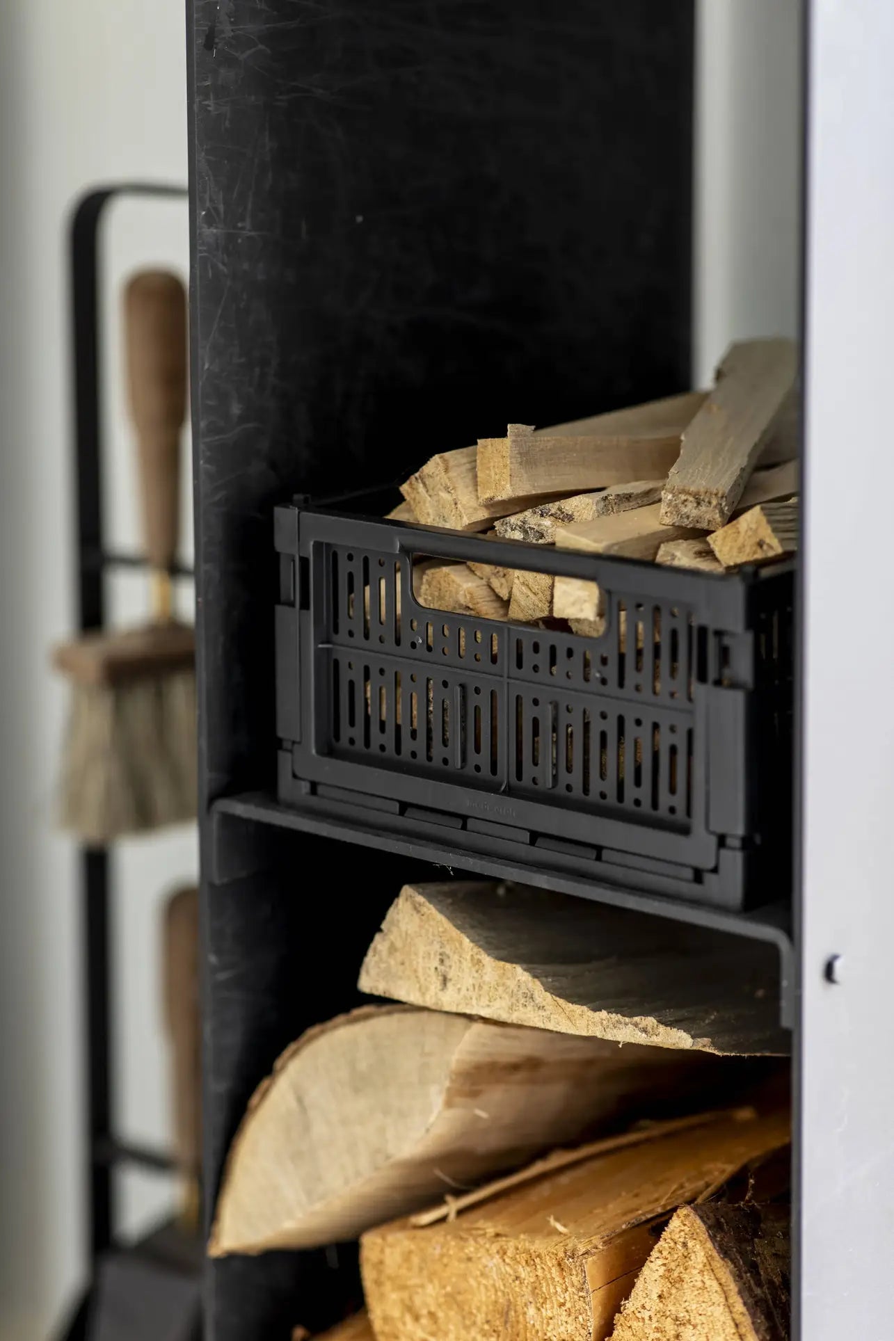 A black shelf holds a Made Crate Midi by Made Crate, an eco-friendly folding crate filled with kindling sticks, while larger firewood logs are stored below. Fireplace tools with wooden handles sit in the background for added convenience.