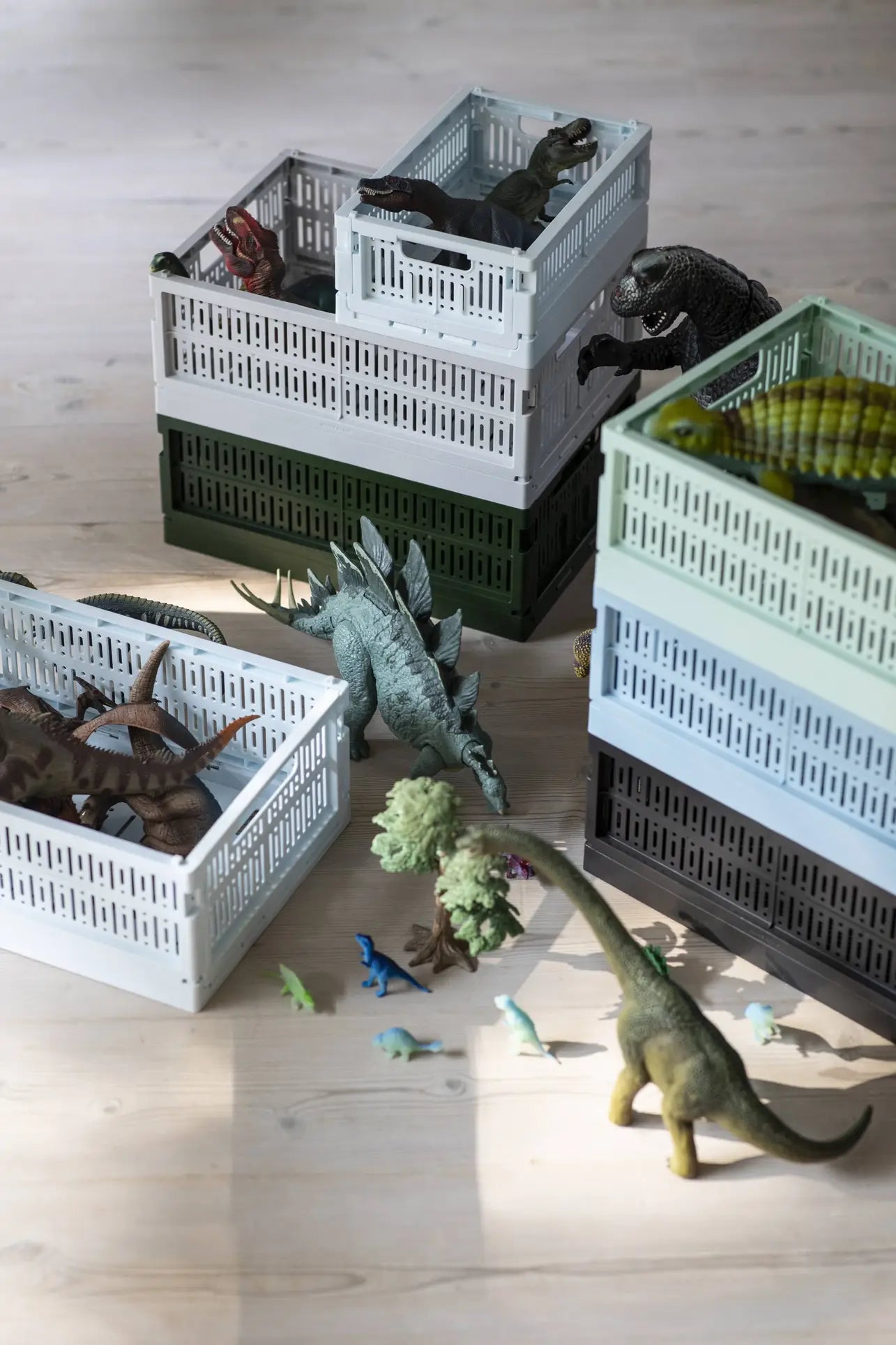 Several toy dinosaurs are arranged in and around Made Crate Midi folding crates by Made Crate, made from recycled plastic. Some dinosaurs sit inside, while others—including a large blue one—highlight the stacking ease on a light wooden floor.