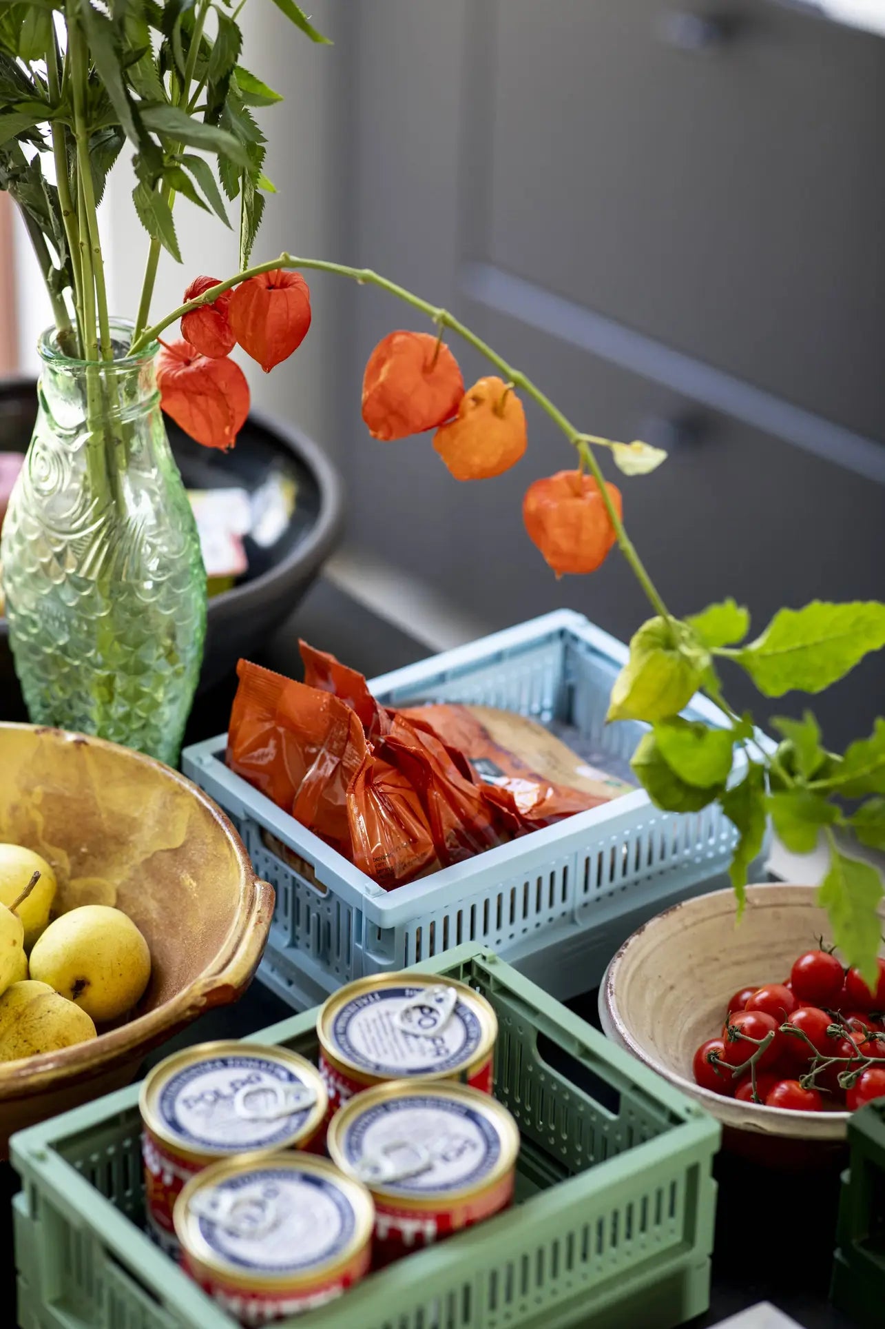 A kitchen counter displays yellow apples, canned goods, cherry tomatoes in a bowl, a vase with orange lantern flowers, and packaged items stored in Made Crate Mini folding crates by Made Crate. A dark cabinet appears in the background.
