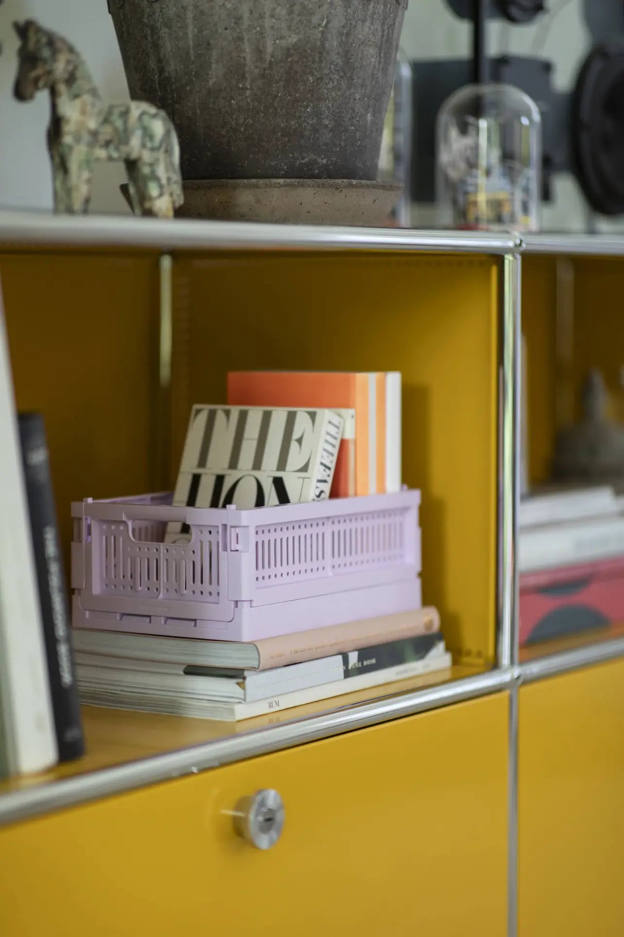 A yellow shelving unit holds books, Made Crate Mini eco-friendly folding crates by Made Crate for stacking, a lavender storage basket, and decorative items like a small horse figurine and a large plant pot on the top shelf.