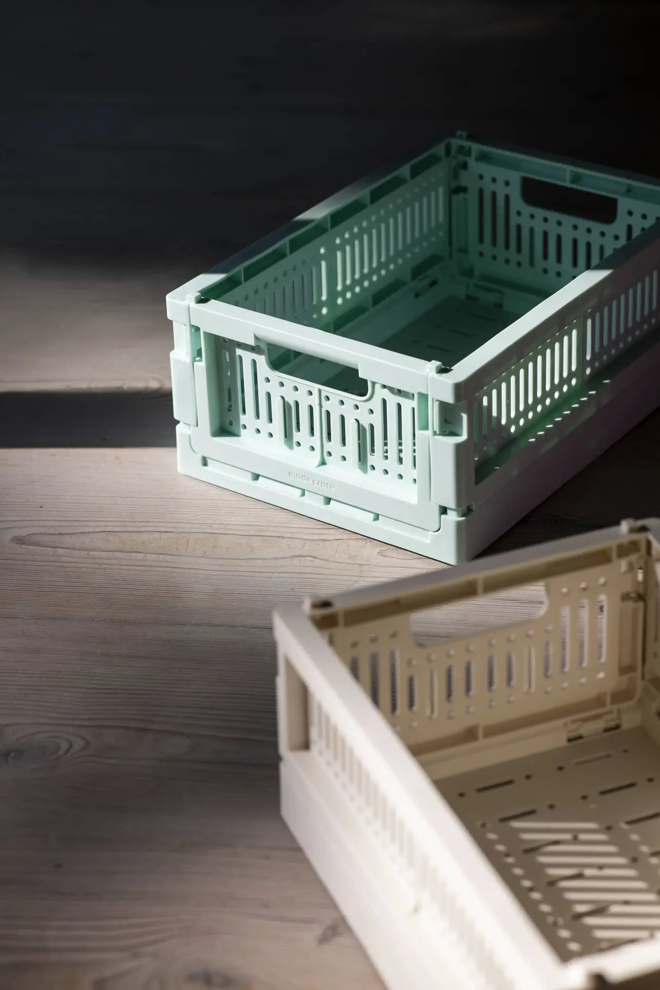 Two Made Crate Mini folding crates by Made Crate—one mint green, one beige—made from recycled plastic, are stacked on a wooden floor with sunlight casting soft shadows, highlighting their compact, eco-friendly stacking convenience.