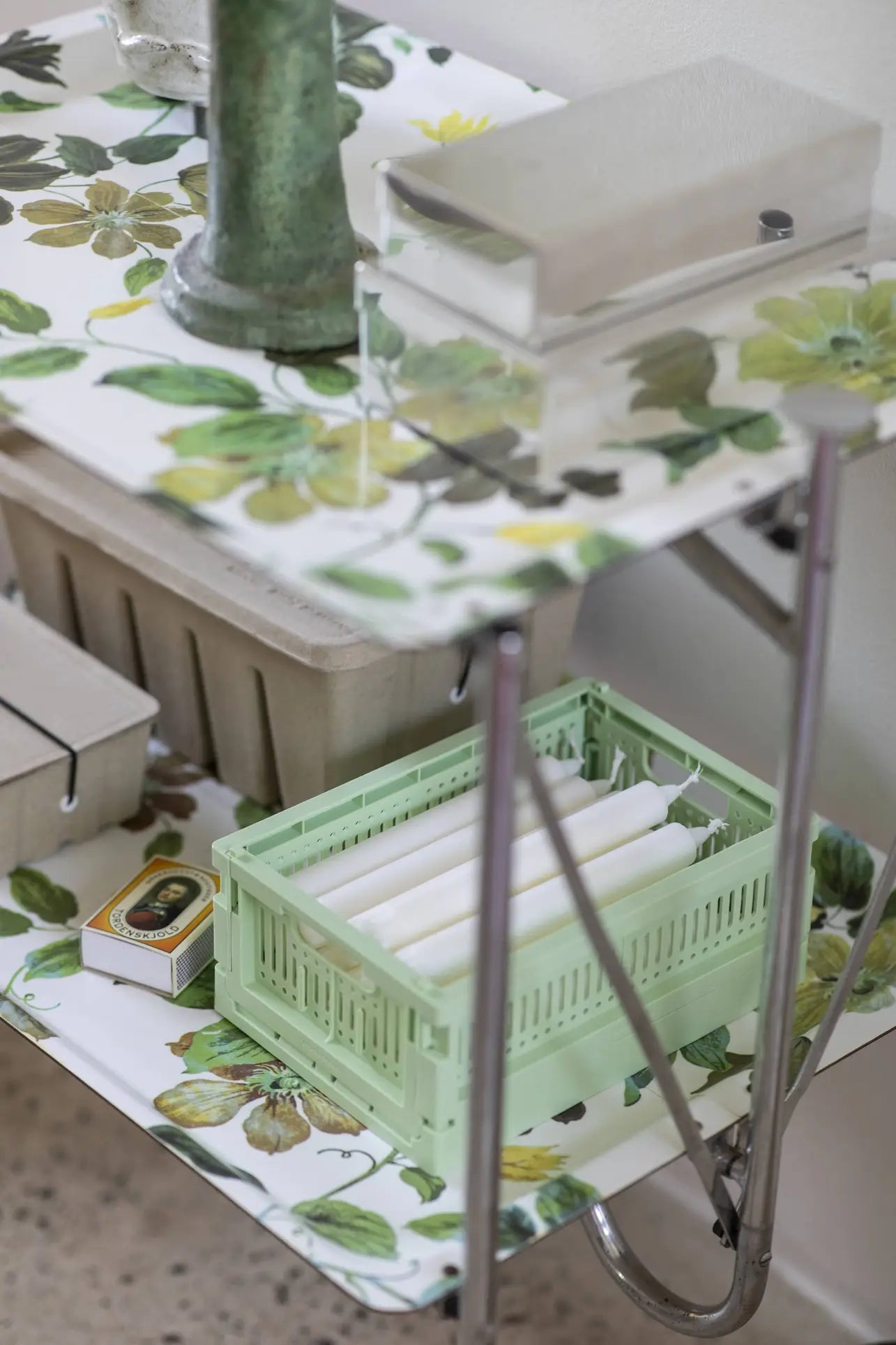 A small cart with a floral-patterned top displays a Made Crate Mini folding crate from Made Crate, filled with white candles, a matchbox, a metal container, and decor. Additional storage boxes are stacked conveniently on the lower shelf.