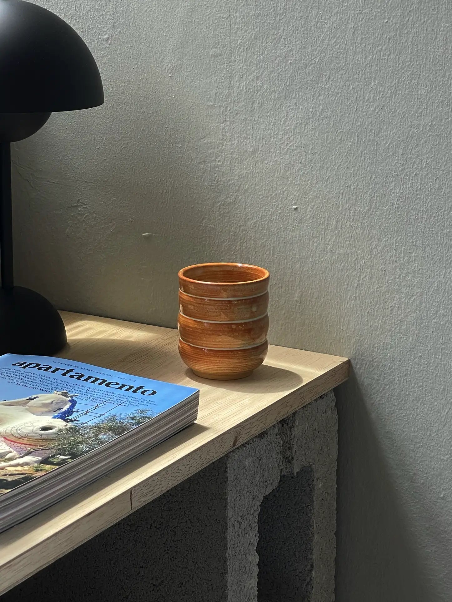 The Mirjam Faber Ceramics Cappuccino Cup Bubble Edition rests on a light wooden table beside a magazine and black lamp, with sunlight casting soft shadows on the textured wall behind.
