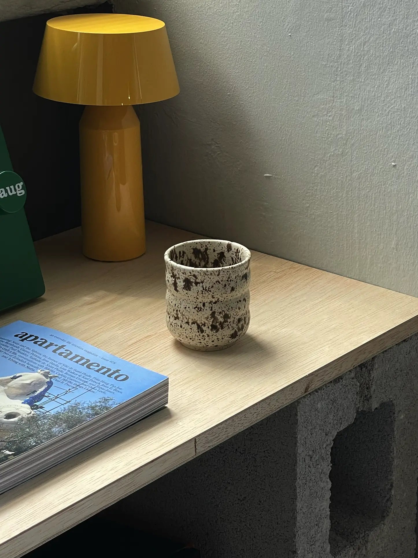 The Mirjam Faber Ceramics Cappuccino Cup Bubble Edition by Mirjam Faber Ceramics rests on a light wooden desk with a concrete base, beside a yellow lamp, green object, and an apartamento magazine with a bird—an elegant Barcelona studio creation.