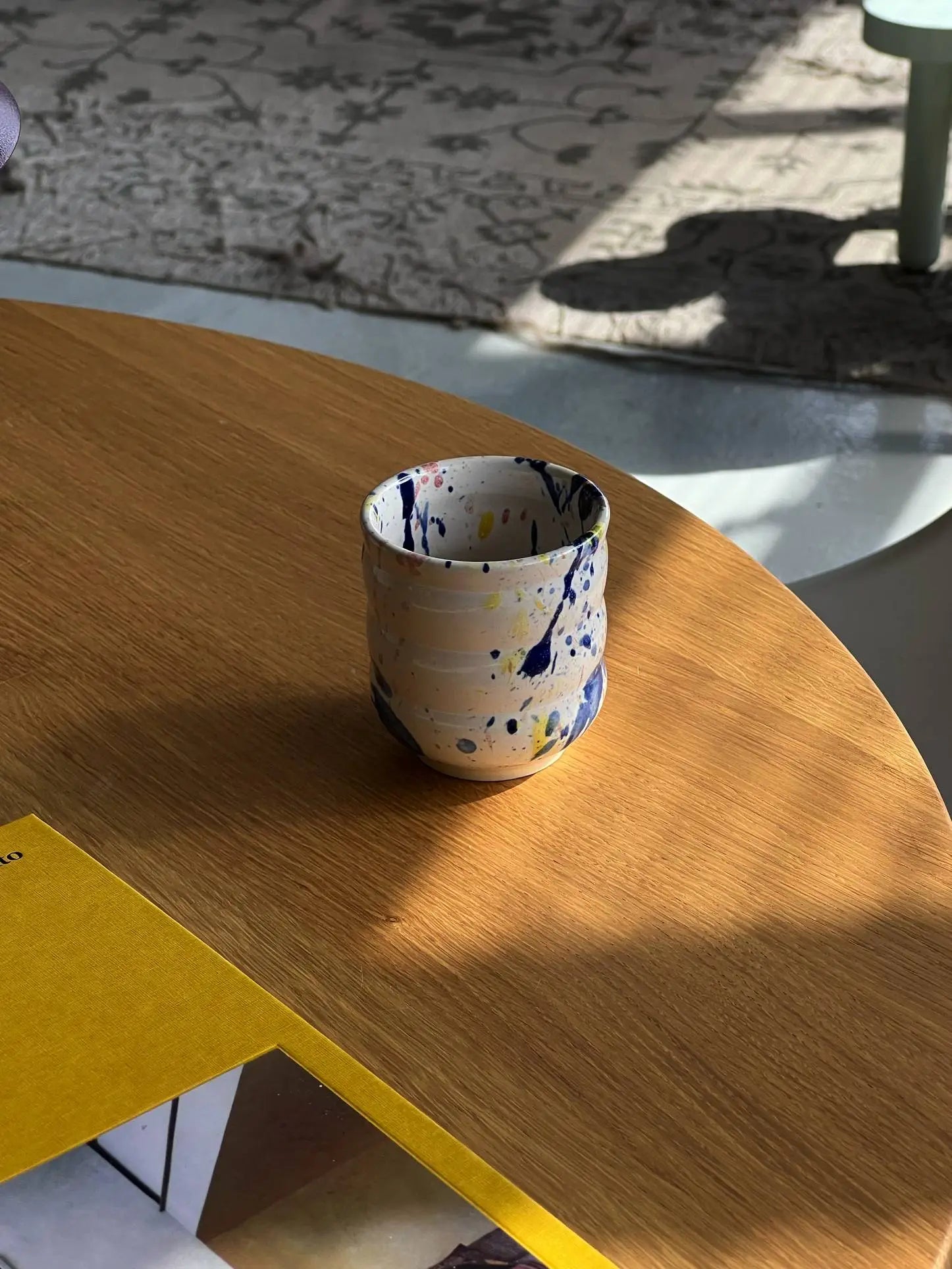 The Mirjam Faber Ceramics Cappuccino Cup Bubble Edition, handmade with blue and yellow splatters, sits on a wooden table beside a yellow book, sunlight casting patterns—an artisanal piece by Mirjam Faber Ceramics of Barcelona.