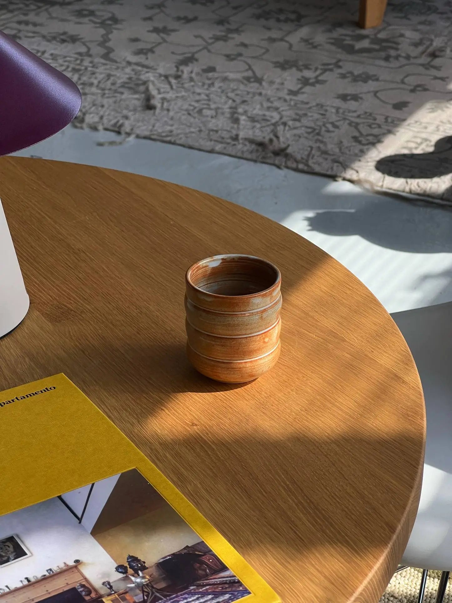 The Mirjam Faber Ceramics Cappuccino Cup Bubble Edition, a small brown handmade cup by Mirjam Faber Ceramics, rests on a wooden table next to a yellow-covered magazine, with sunlight and a patterned rug in the background.