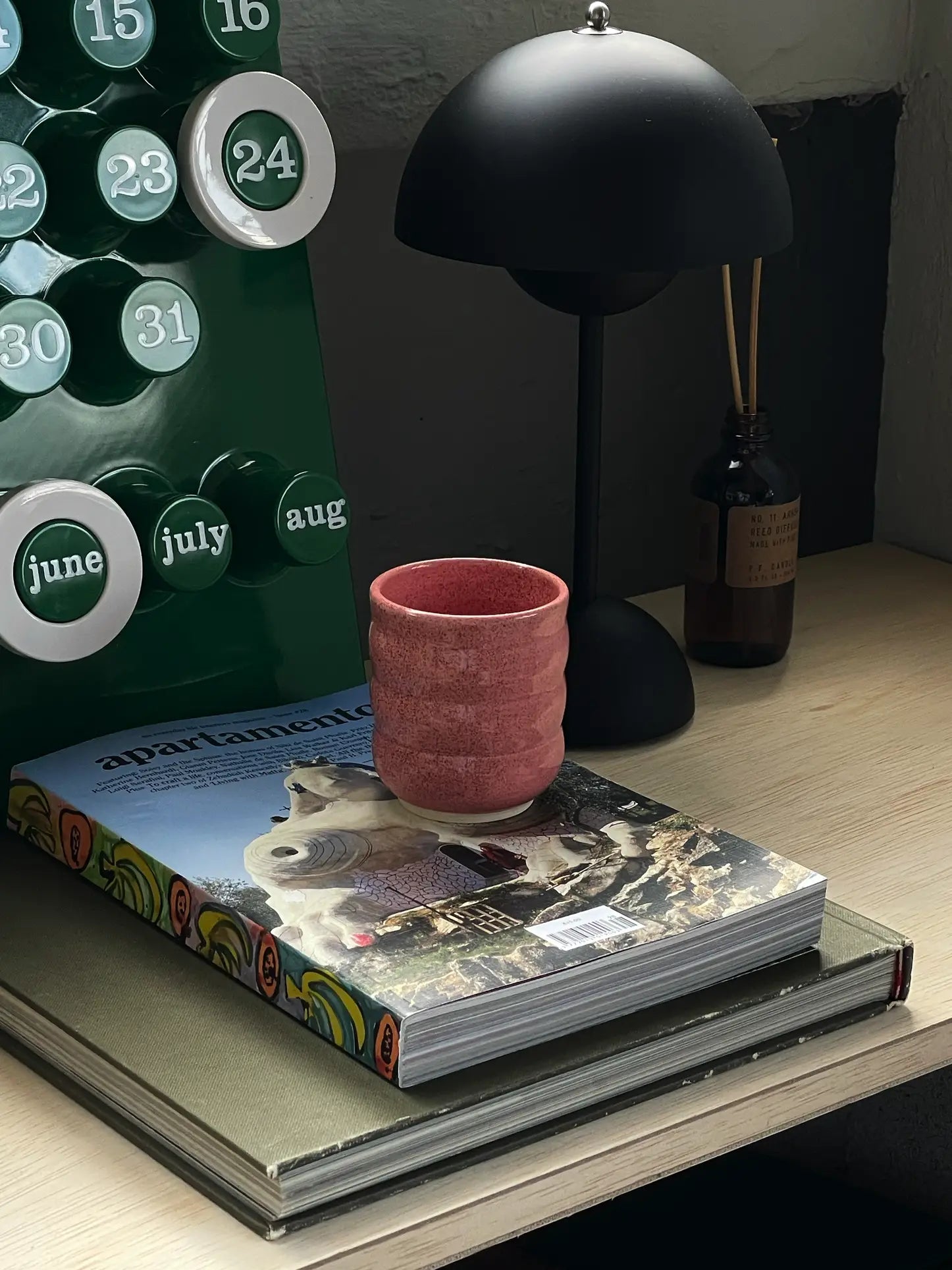 The Mirjam Faber Ceramics Cappuccino Cup Bubble Edition in pink rests on an illustrated book next to a calendar showing June–August, with a black desk lamp and diffuser on a wooden desk—echoing Barcelona studio charm.