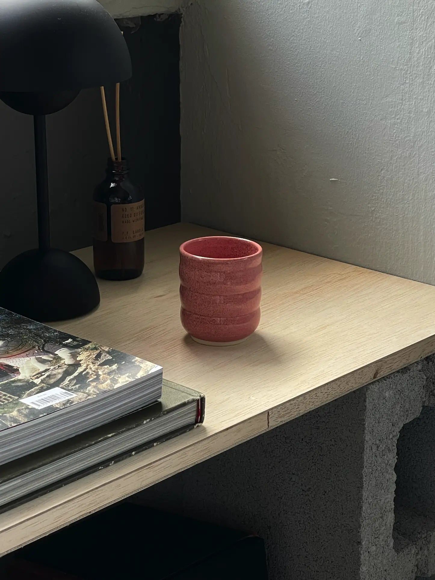 The Mirjam Faber Ceramics Cappuccino Cup Bubble Edition, in pink ceramic with a bubble design, sits on a light wooden desk beside books, a dark lamp, and reed diffusers—crafted by Mirjam Faber Ceramics in Barcelona.