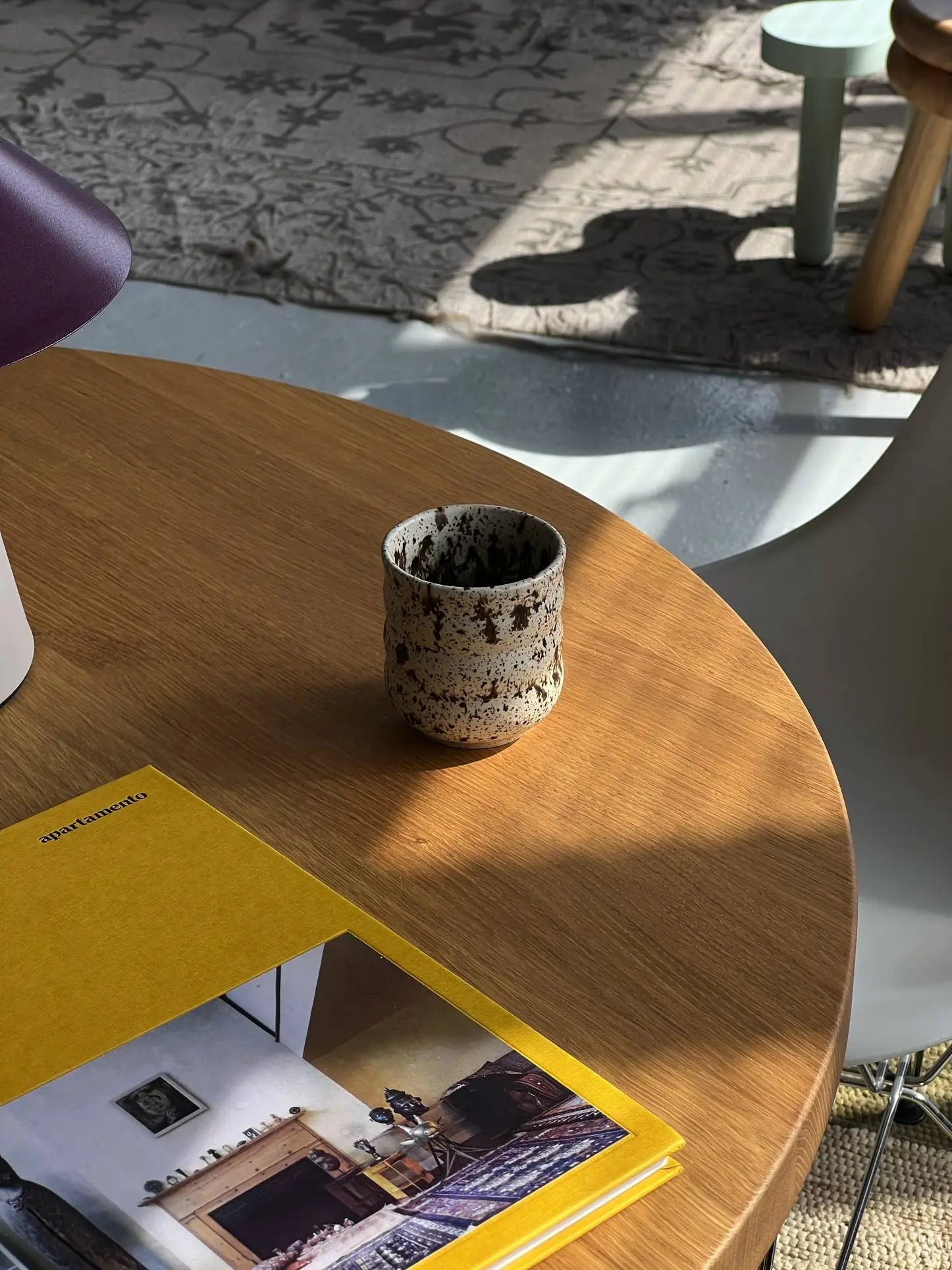 The Mirjam Faber Ceramics Cappuccino Cup Bubble Edition by Mirjam Faber Ceramics sits on a round wooden table next to a yellow-covered magazine, sunlight and shadows dancing across the surface and a patterned rug in the background.