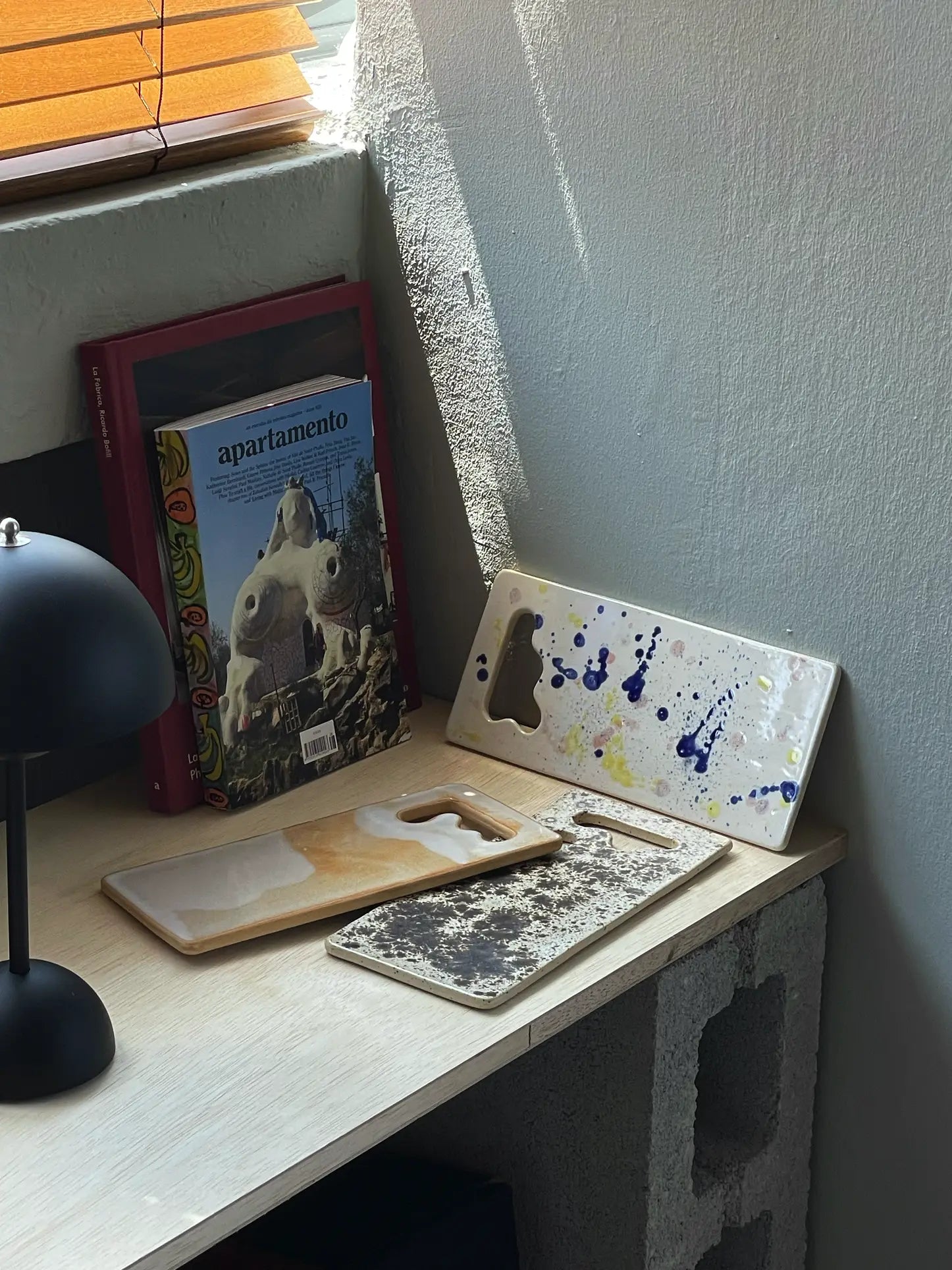 A small desk with a black lamp, books including Apartamento, and Mirjam Faber Ceramics Sushi Plates from Mirjam Faber Ceramics, featuring unique cutouts, set against a wall with a concrete block and wooden blinds.