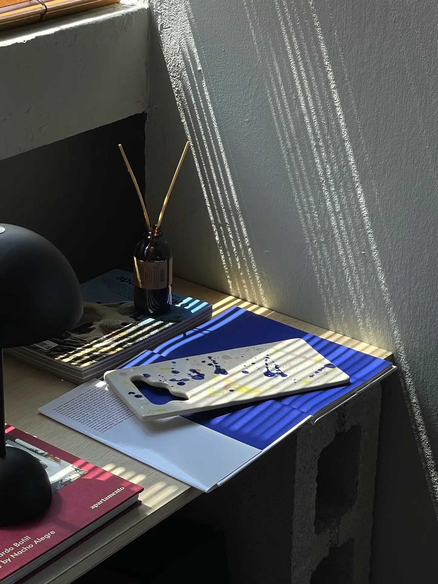 A desk by a sunlit window with books, a blue notebook, a Mirjam Faber Ceramics Sushi Plate featuring unique cutouts, a dark lamp, and a brown reed diffuser bottle casting shadows on the wall.